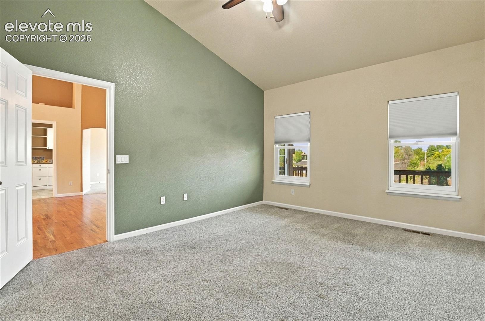 Room with a vaulted ceiling, neutral wall coloring, carpeted flooring, two windows with integrated shades, and an attached ceiling fan with lighting