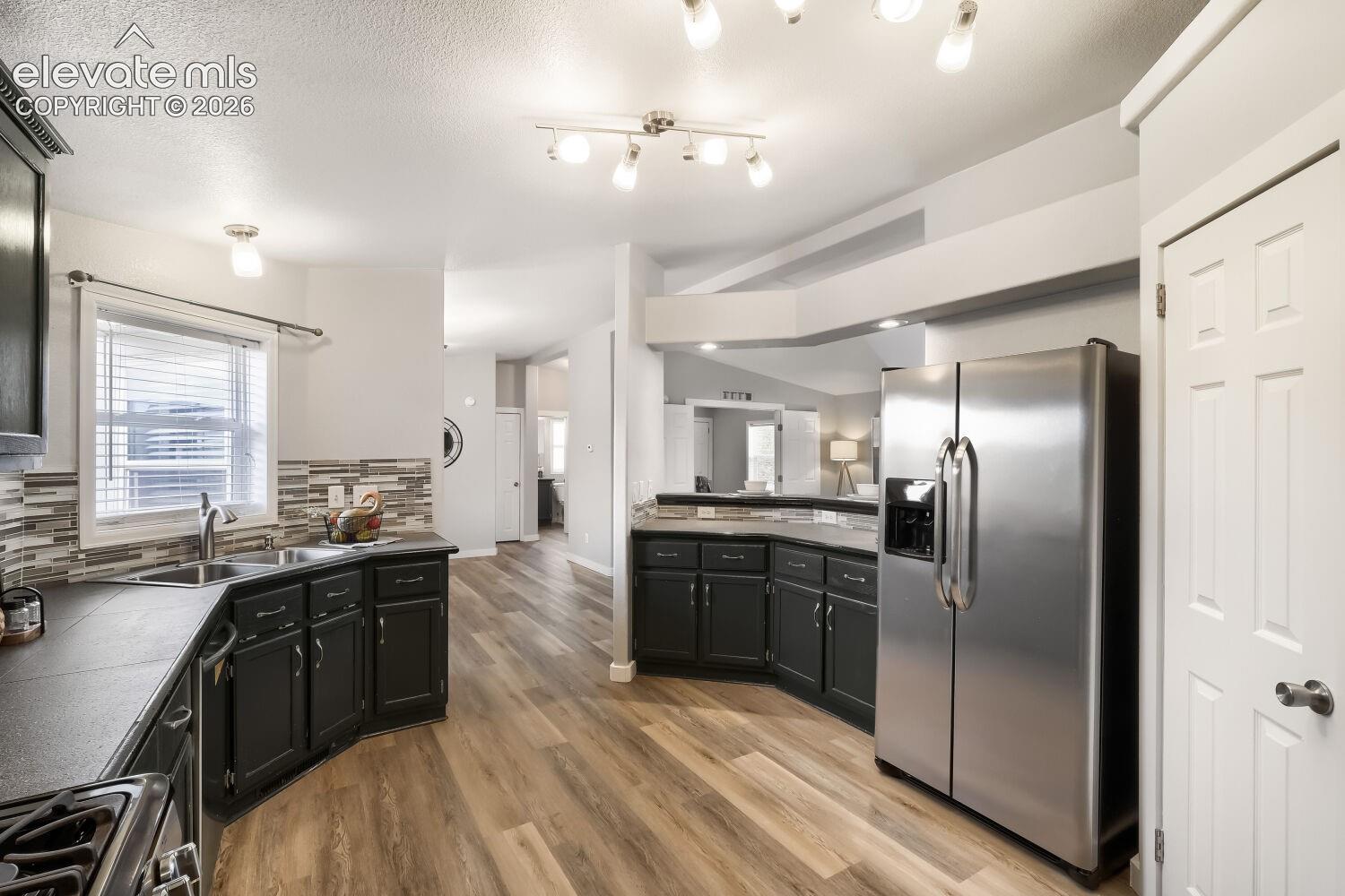 Kitchen featuring wood-finish flooring, dark cabinetry, stainless steel side-by-side refrigerator, multi-toned tile backsplash, and multi-light track lighting
