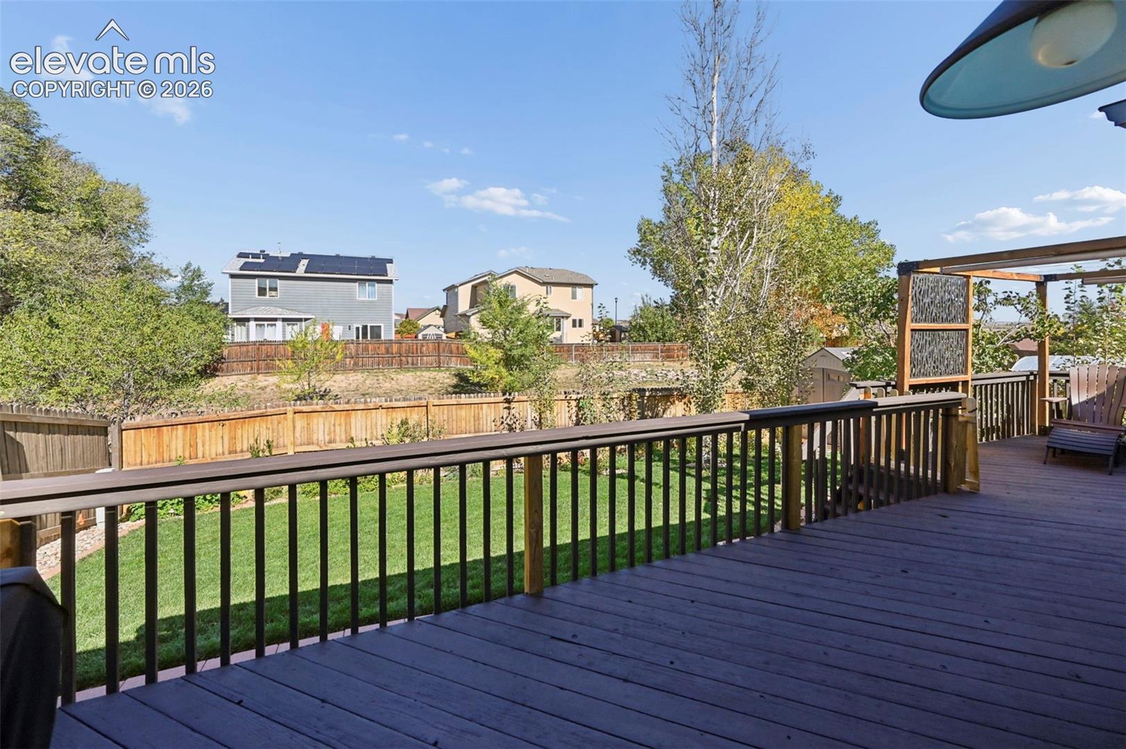 Elevated wooden deck featuring a horizontal railing, a pergola structure with lattice paneling, and an expansive lawn enclosed by a wooden privacy fence