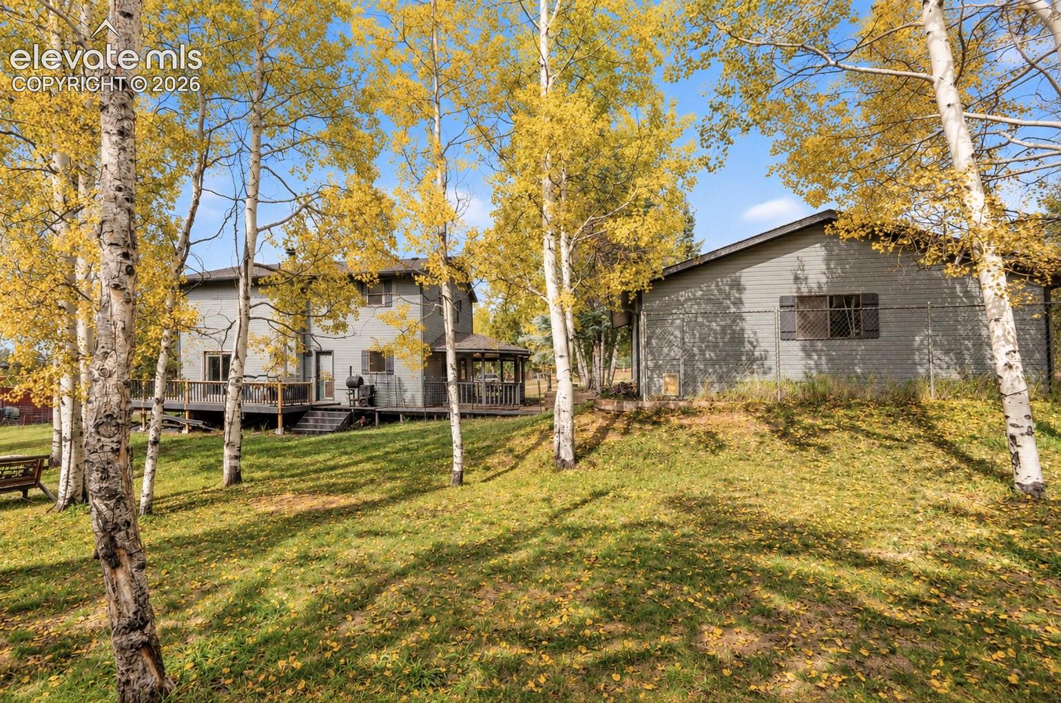 Expansive green lawn with mature trees, a two-story residence with a multi-level deck, and an additional structure featuring horizontal siding and dark shutters