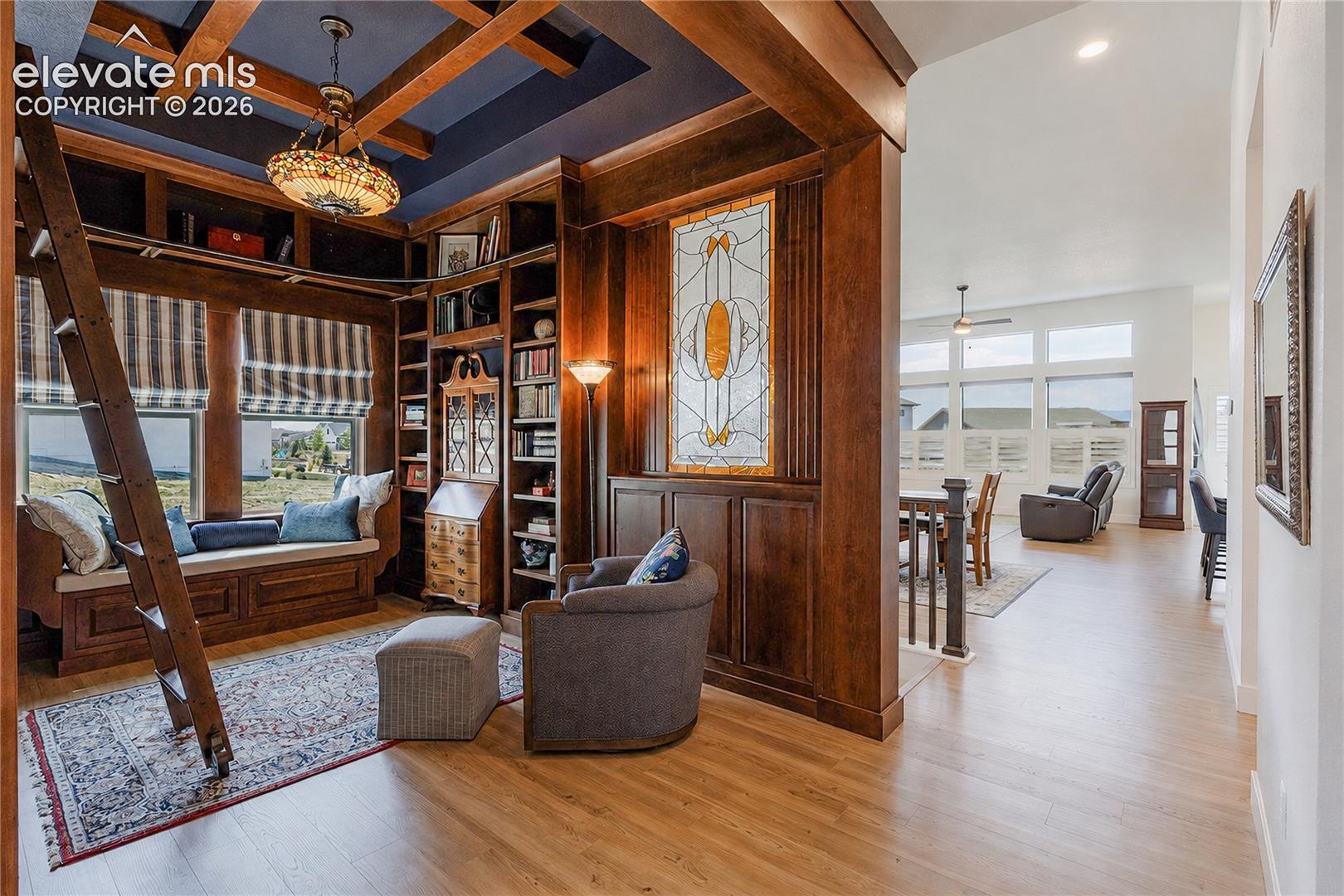 Custom built-in bookcases with a rolling ladder, coffered ceiling with a central pendant fixture, and a built-in window seat