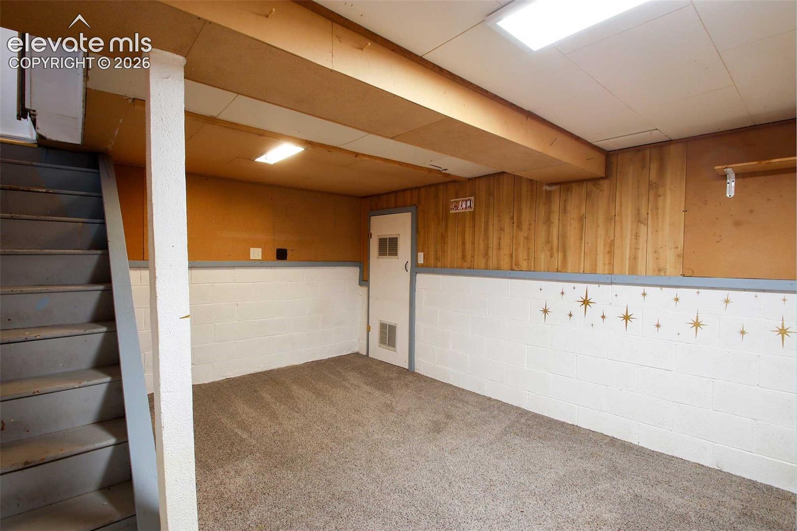 Carpeted flooring, painted cinder block walls, wood paneling, stair access, and overhead lighting
