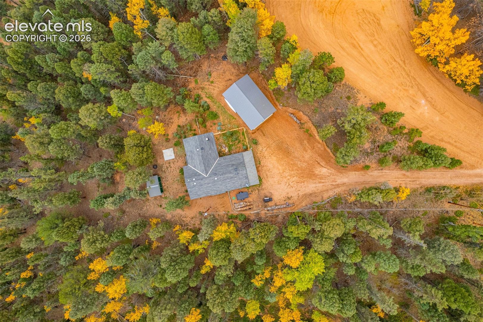 Aerial perspective of a multi-structure property featuring a main residence and detached garage, situated within a wooded lot characterized by mature coniferous and deciduous trees and earthen access paths