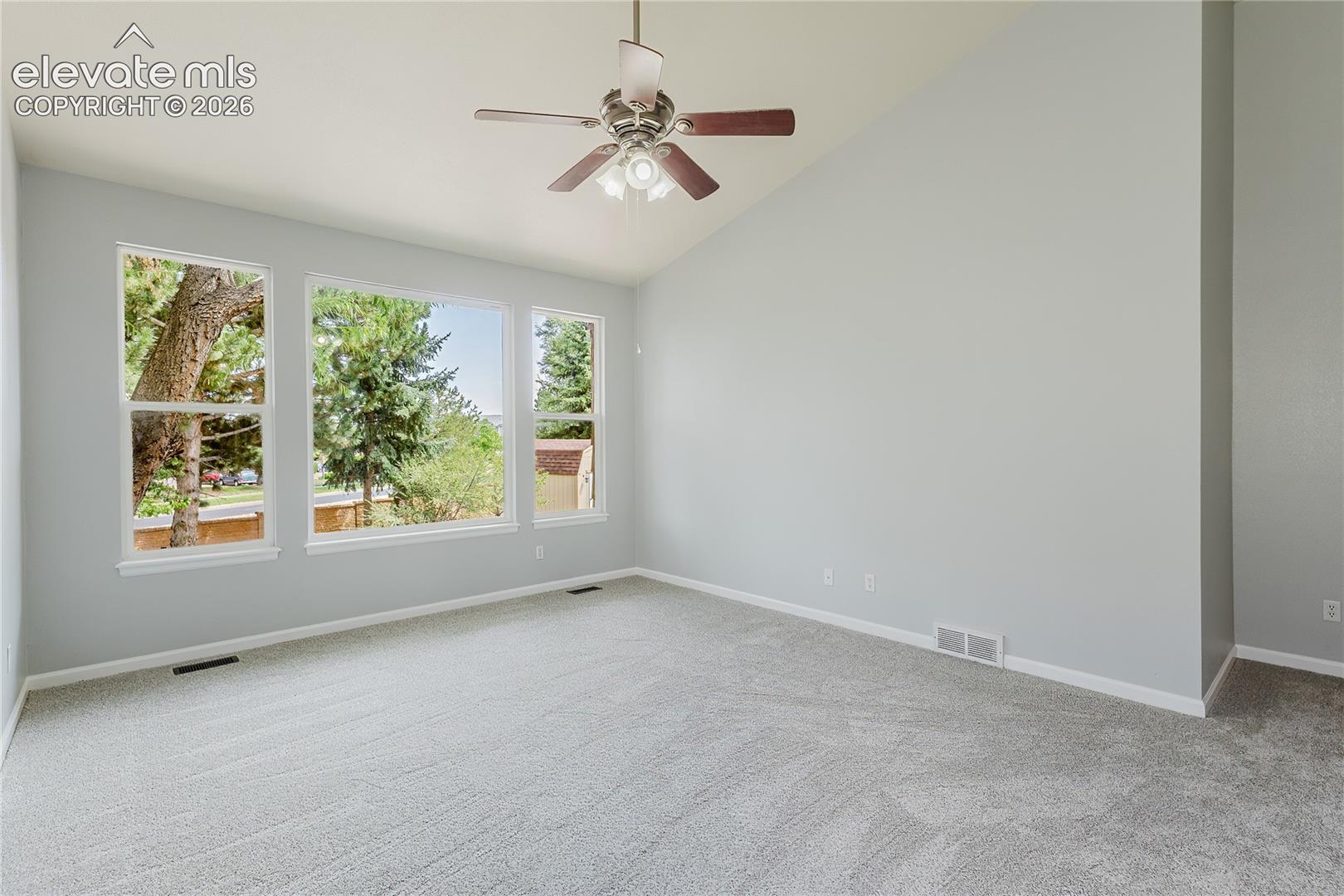 Vaulted ceiling, ceiling fan with light fixture, light gray walls, light-toned carpet, and a large window array