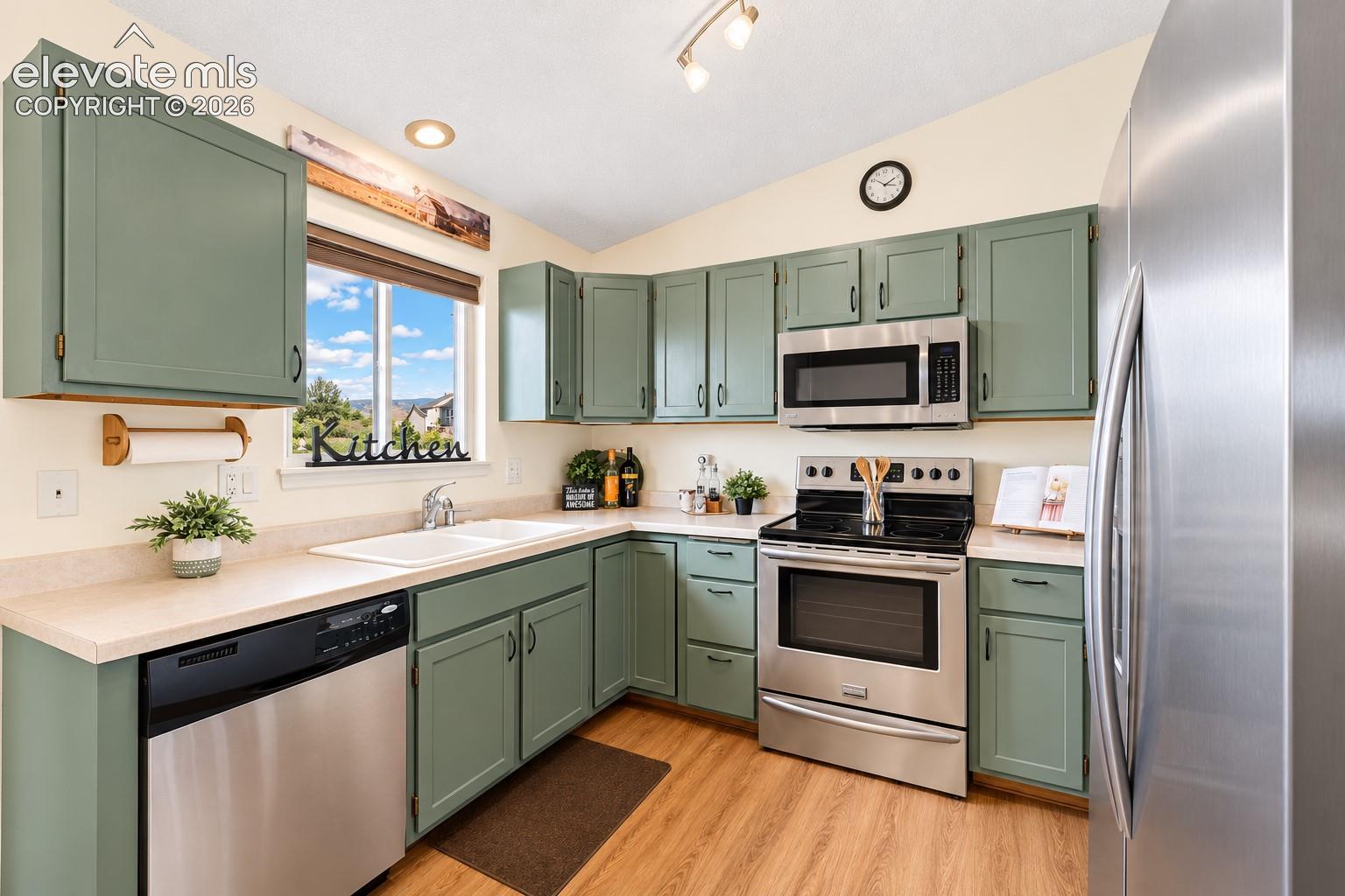 Kitchen featuring sage green shaker-style cabinetry, stainless steel appliances, dual-basin sink, solid-surface countertops, and wood-finish flooring