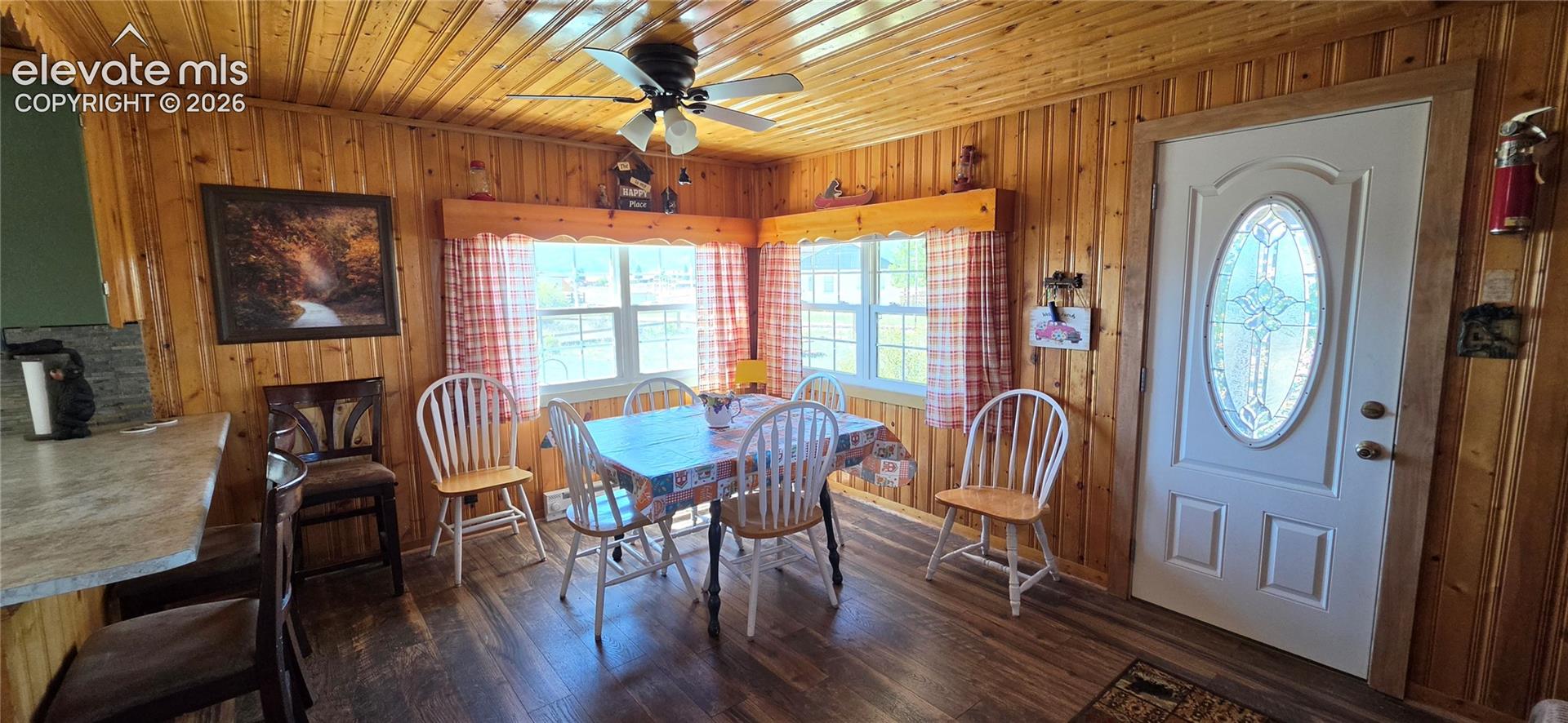 Wood-paneled walls and ceiling, wood-finish flooring, two large windows, white entry door with oval decorative glass, and a ceiling fan with integrated lighting