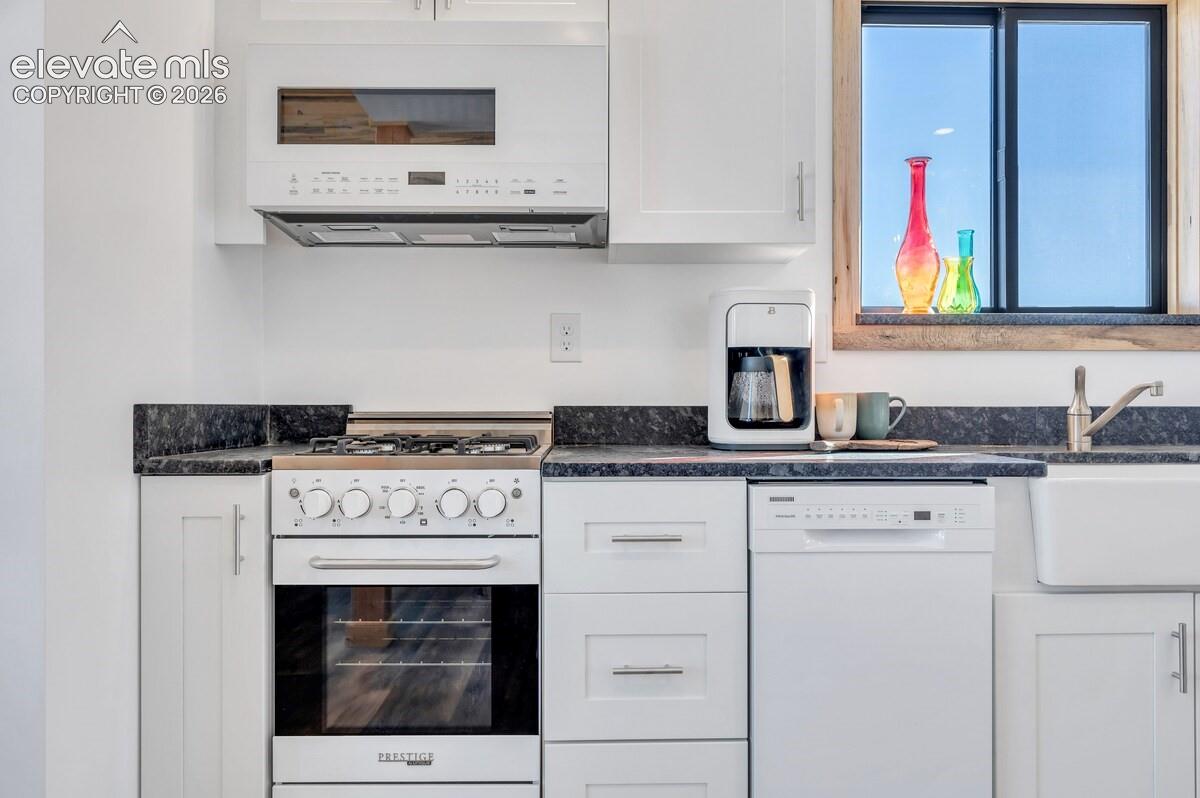 Kitchen featuring white shaker-style cabinetry, dark stone-finish countertops, apron-front farmhouse sink, gas range, and an integrated over-the-range microwave