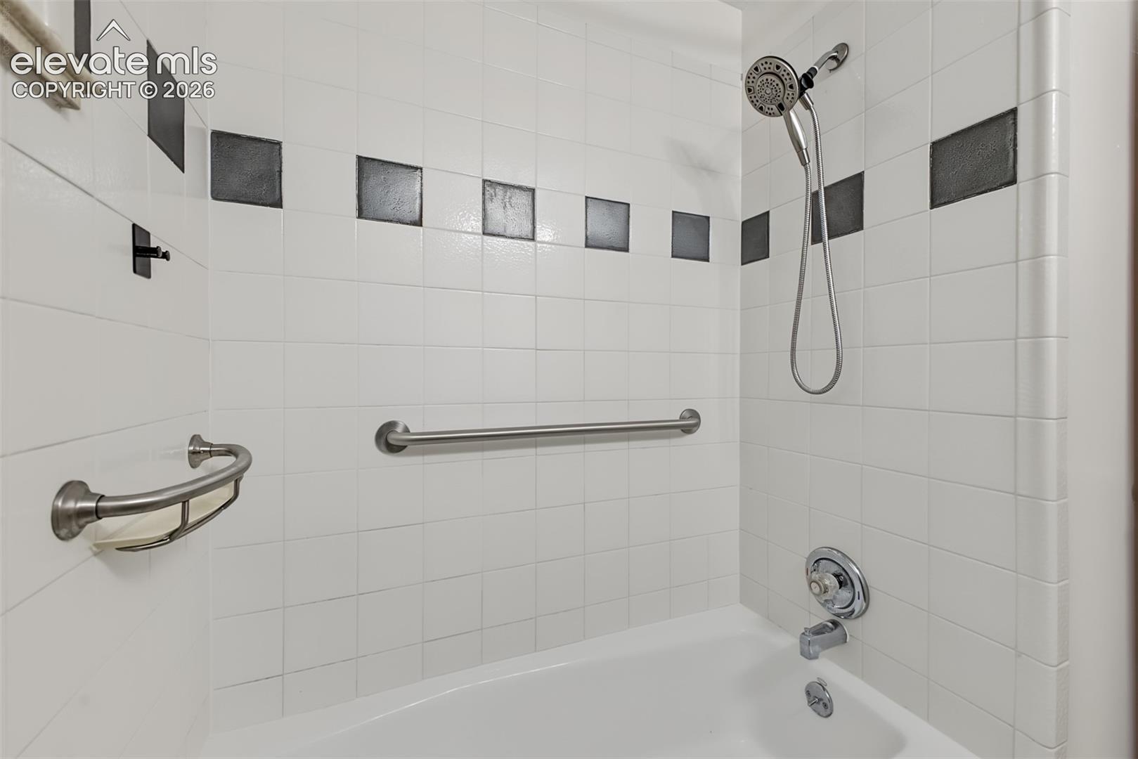 Bathtub alcove featuring white wall tiling with black accent tiles, a polished chrome hand-held shower fixture, and stainless steel grab bars