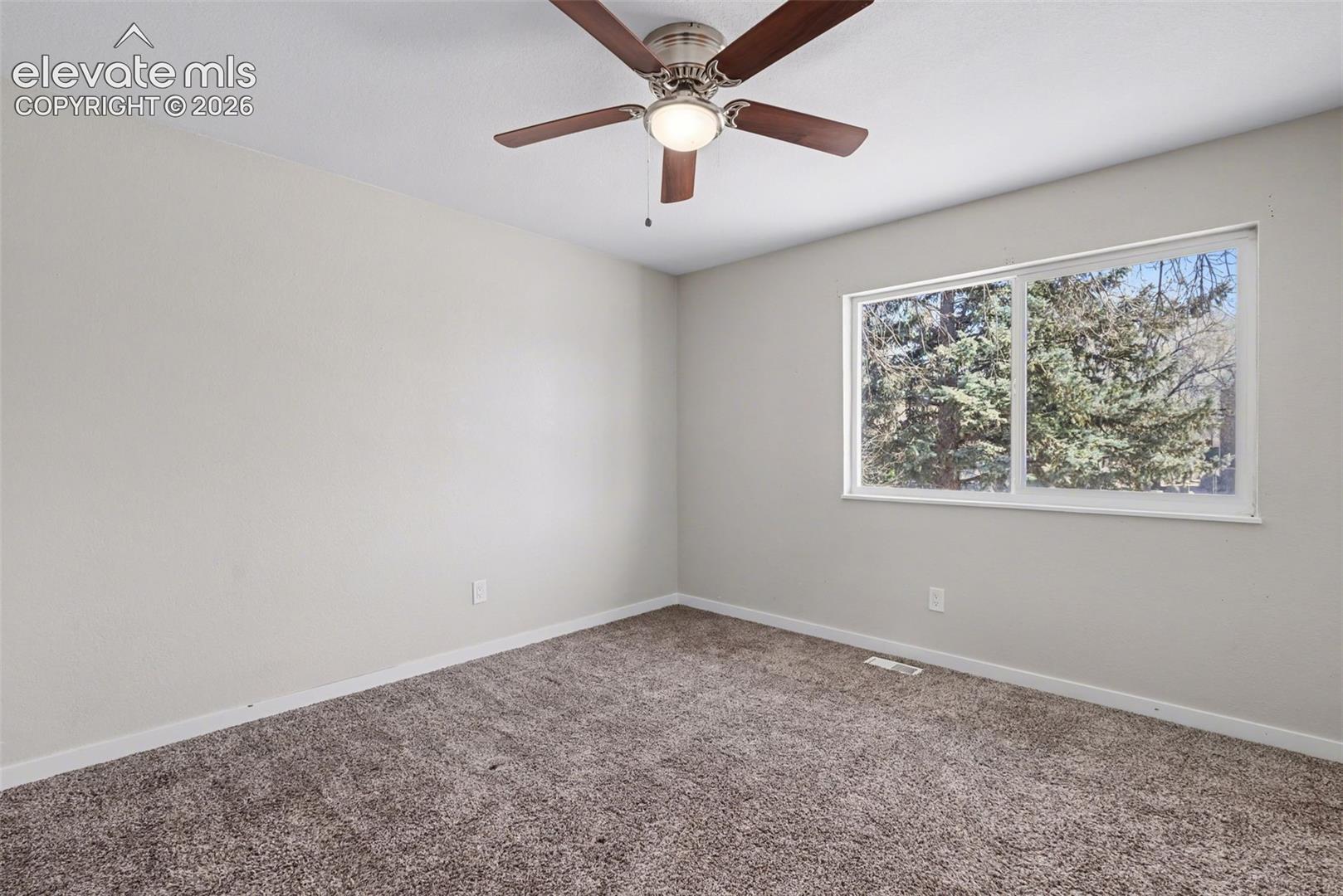 Neutral wall tones, carpeted flooring, sliding window with exterior tree greenery, and a ceiling-mounted light fixture with a five-blade fan