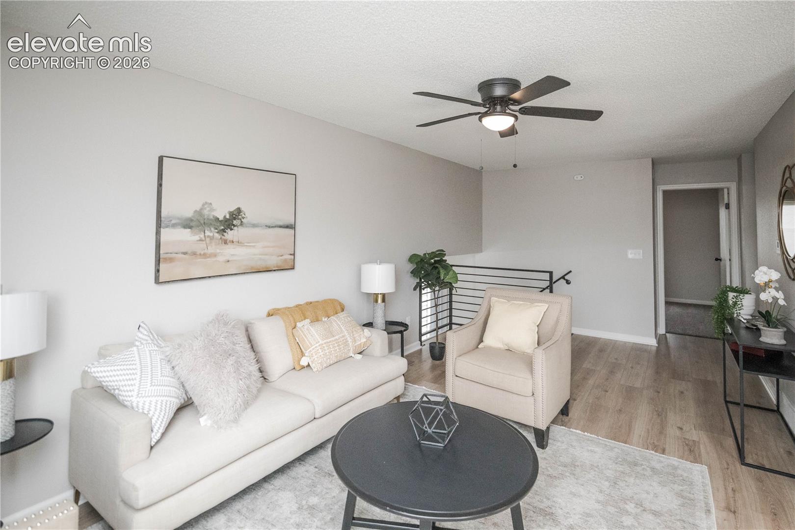 Living area featuring wood-finish flooring, a matte black ceiling fan with integrated lighting, and a metal horizontal-bar railing