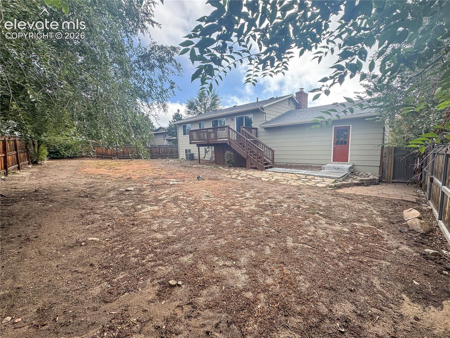 Backyard featuring a raised wooden deck, a stone patio, mature trees, and perimeter wood fencing