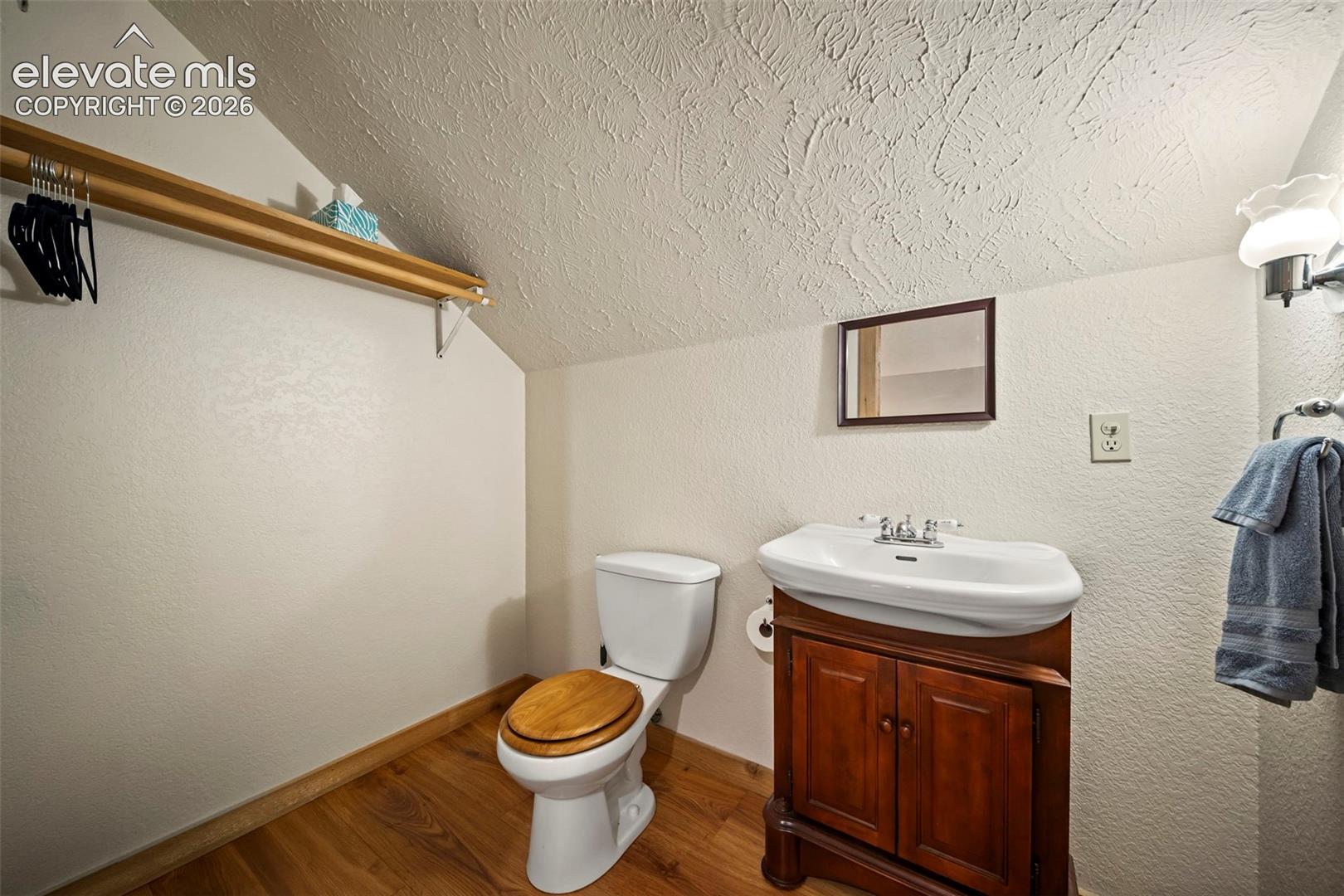 Bathroom featuring wood-finish flooring, a wall-mounted wooden shelf, a vanity with a white porcelain sink and dual-handle faucet, and a wall-mounted light fixture