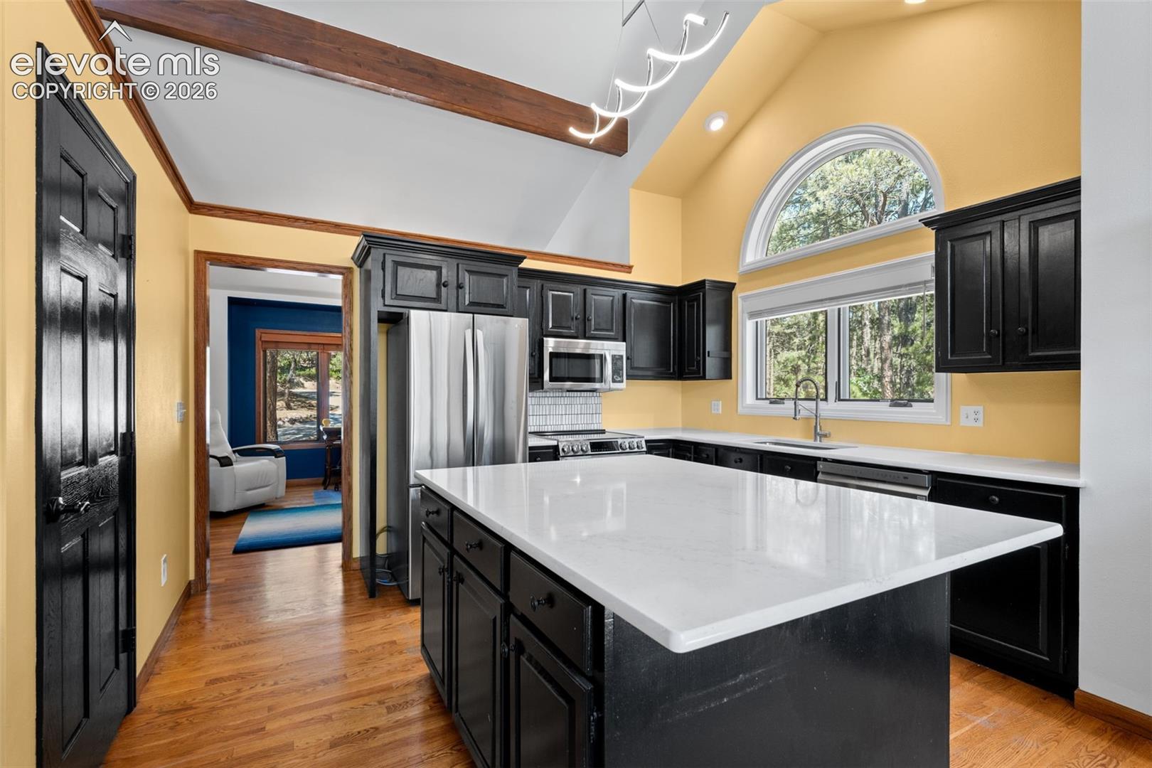 Kitchen featuring vaulted ceilings with exposed wood beams, a large arched window, white countertops, black cabinetry, and stainless steel appliances