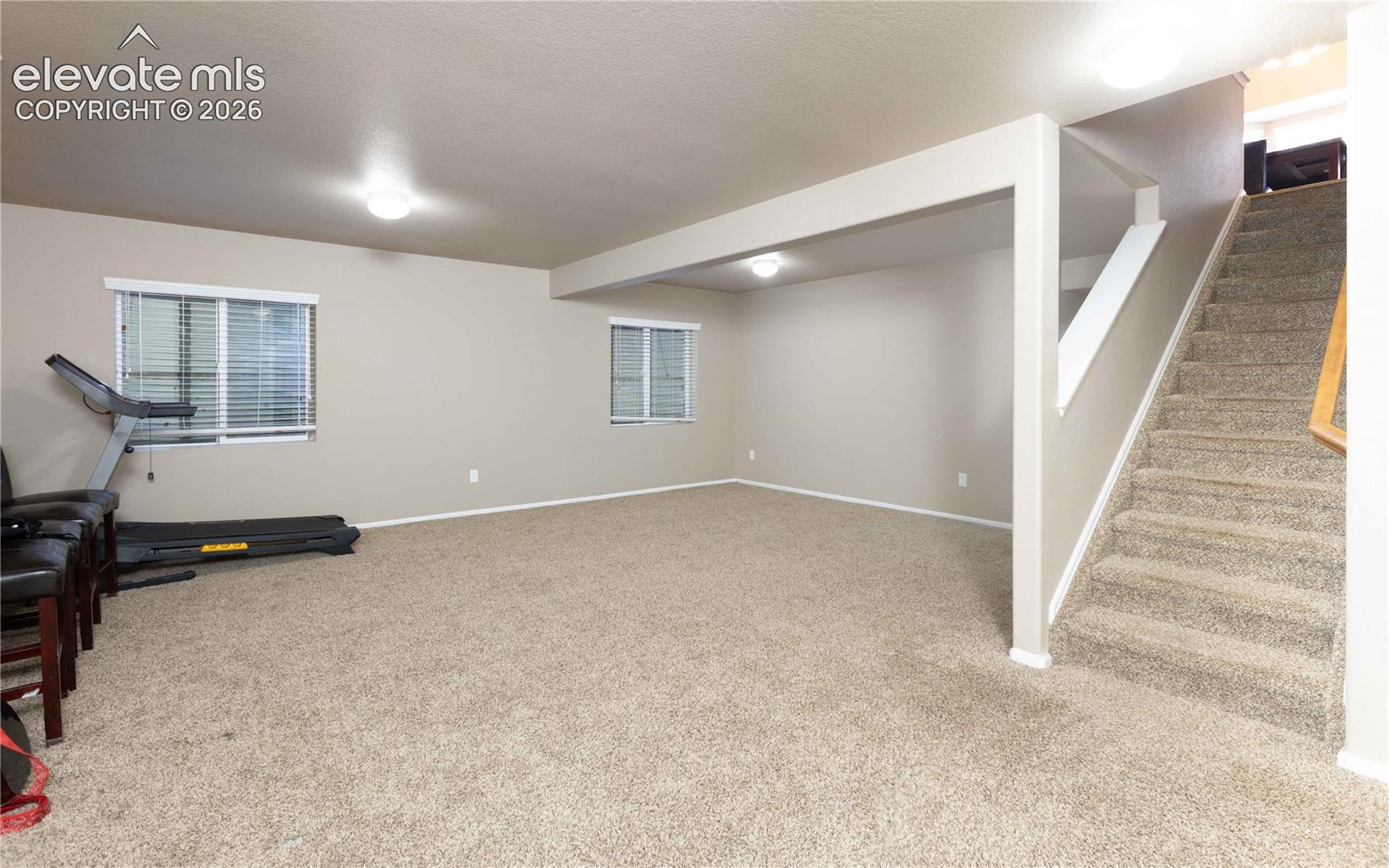 Open-concept interior space featuring neutral wall tones, carpeted flooring, two horizontal sliding windows with blinds, and a staircase with a wooden handrail