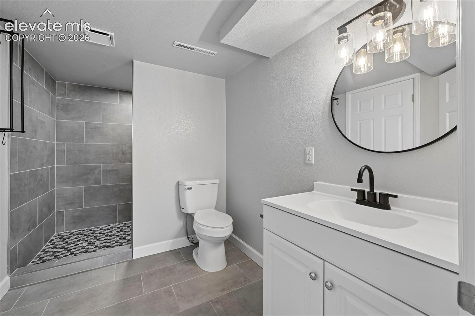Basesment Bathroom down the hall that is also ensuite with a secondary suite in the basement!  featuring a walk-in shower with grey tile surround and tile floor, vanity with white countertop and integrated sink, matte black hardware, and a circular wall-mounted mirror with integrated lighting