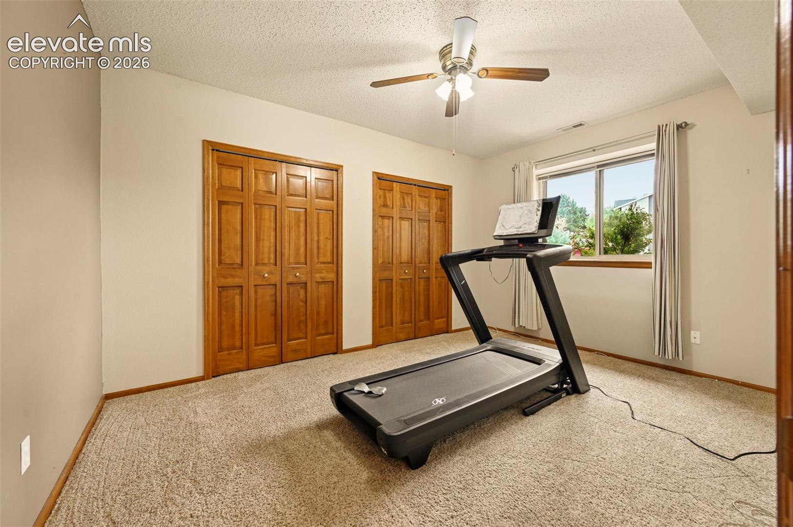 Basement bedroom with neutral wall paint, two wood-finish bi-fold closet doors, carpeted flooring, and a ceiling fan with light fixture