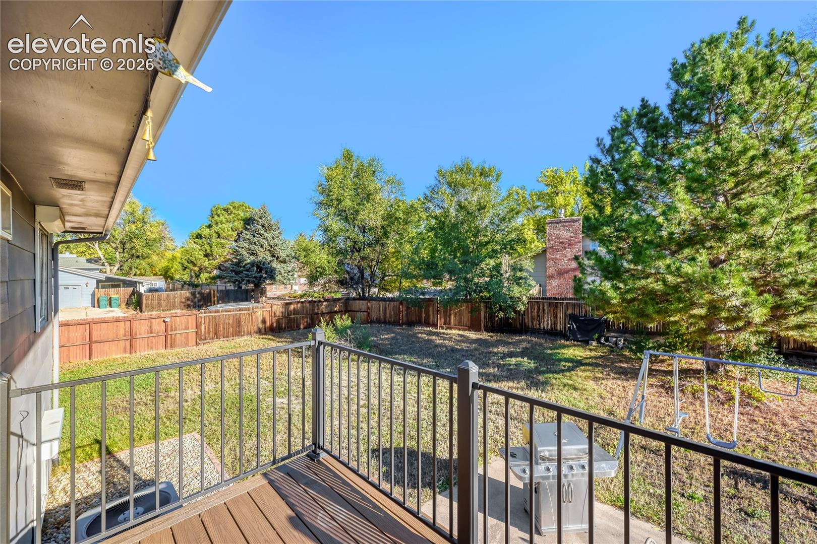 Elevated deck featuring composite decking and black metal railing, overlooking a fenced backyard with mature trees and a concrete patio