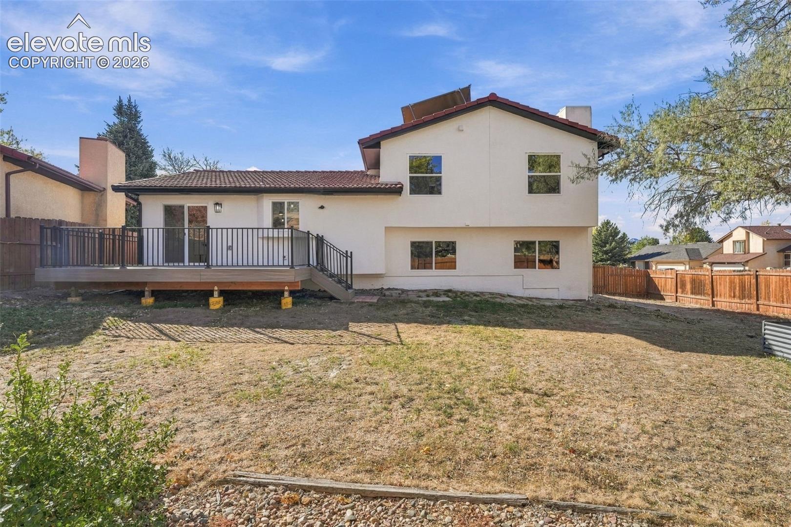 Two-story exterior featuring light-colored stucco siding, a multi-pitched tile roof, an elevated deck with black metal railing, and enclosed wood fencing