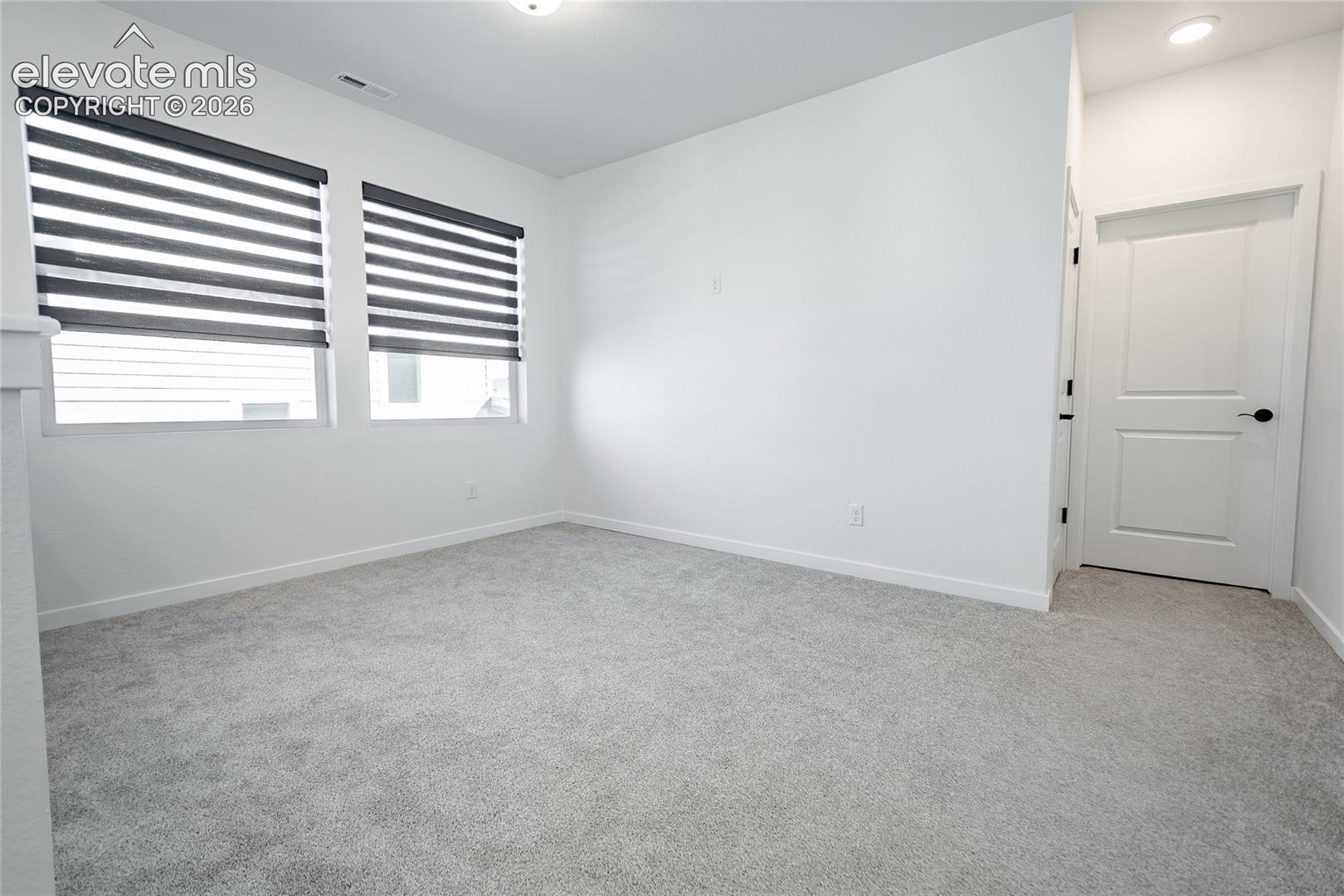 Neutral-toned carpeted flooring, dual windows with fitted light-filtering shades, recessed ceiling lighting, and a multi-panel interior door with matte black hardware