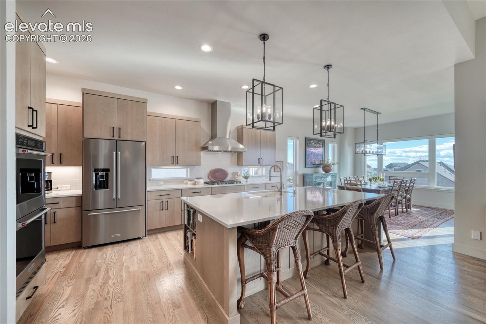 Kitchen featuring light wood-finish cabinetry, a quartz-topped center island with integrated seating, stainless steel appliances, and geometric pendant lighting over wood-finish flooring