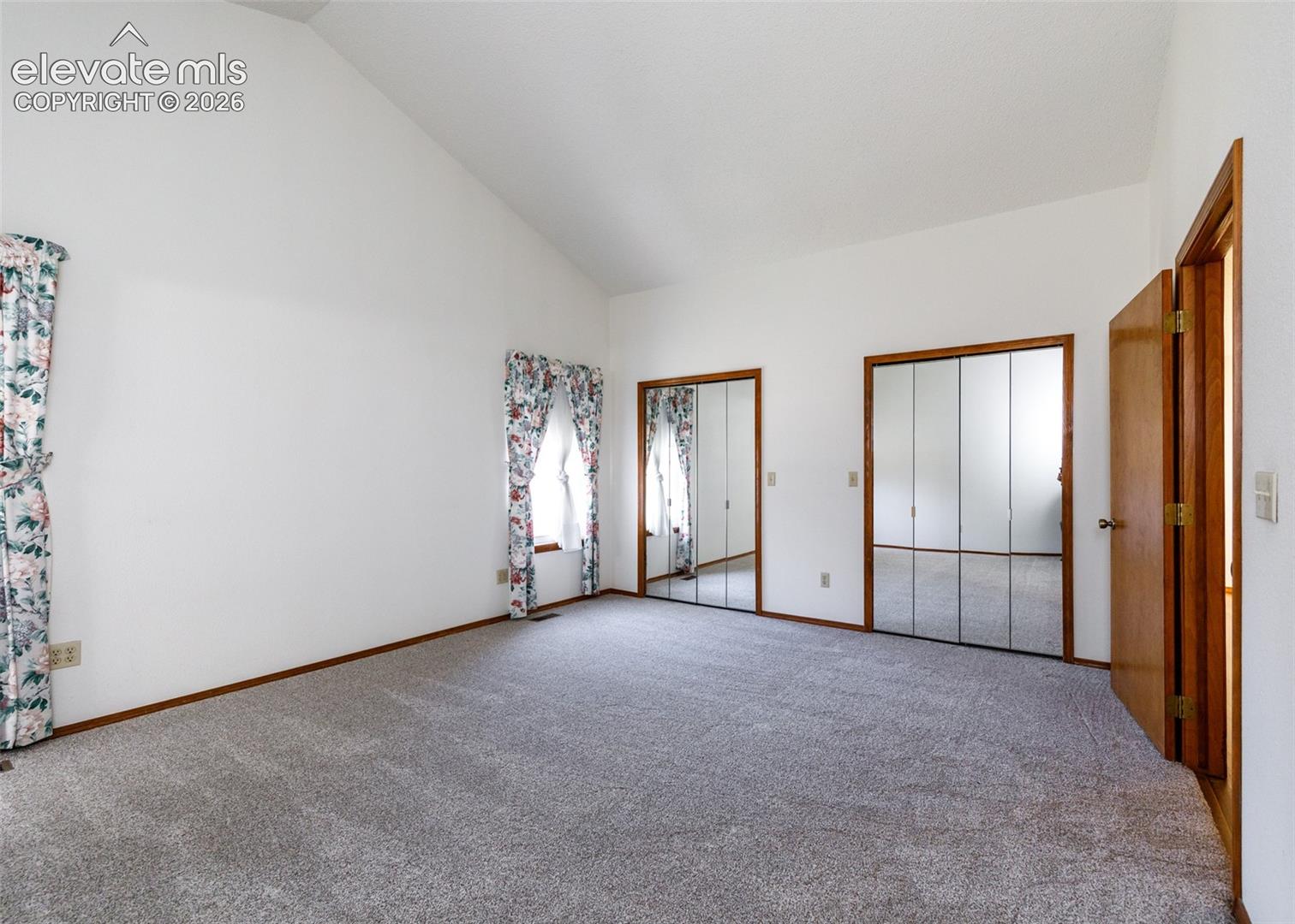 Vaulted ceiling, two mirrored closet doors, two windows, and gray carpet