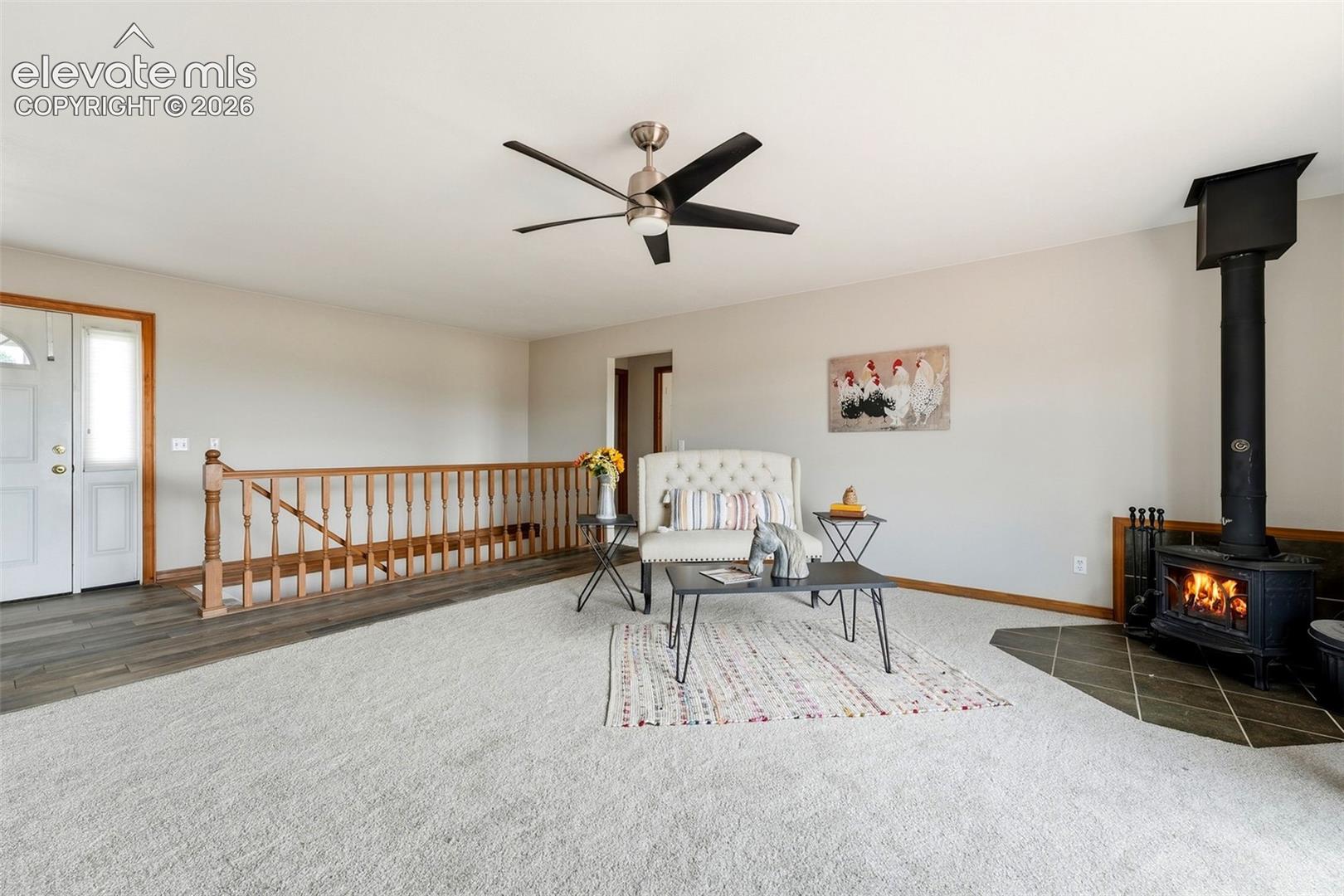 Carpeted living space featuring a wood-burning stove with a tiled hearth, ceiling fan, wood-finish flooring, and a wooden staircase railing