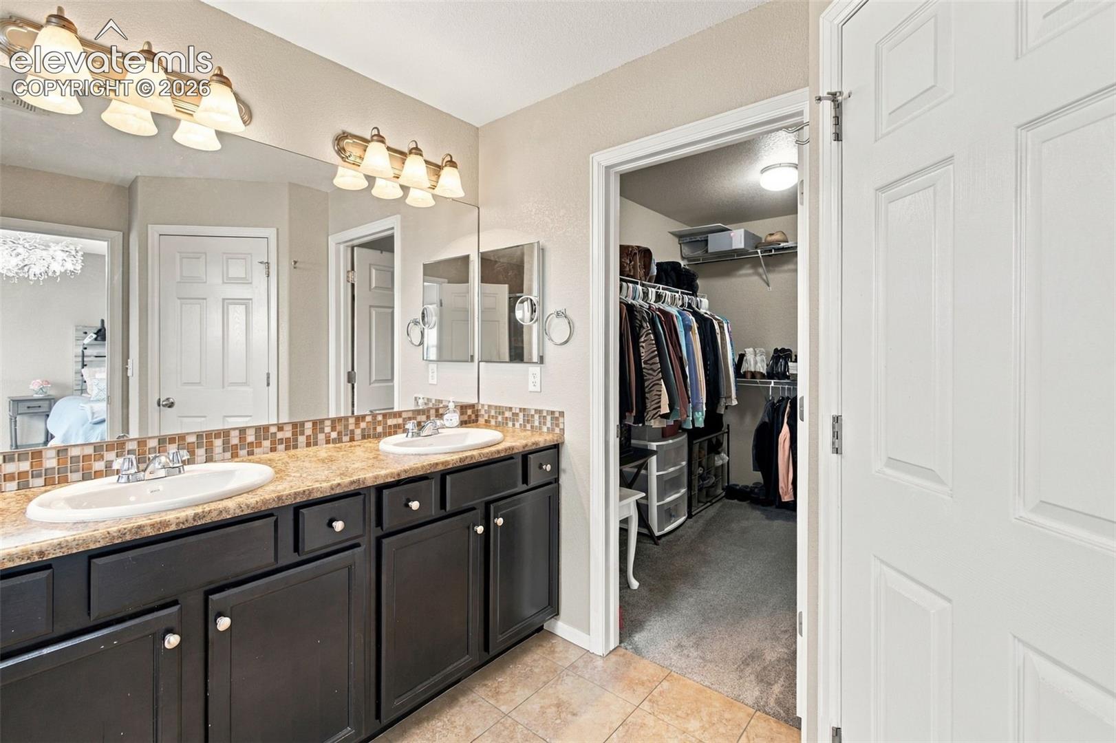 Dual vanity featuring stone-finish countertops, tile backsplash, dark cabinetry, and dual light fixtures