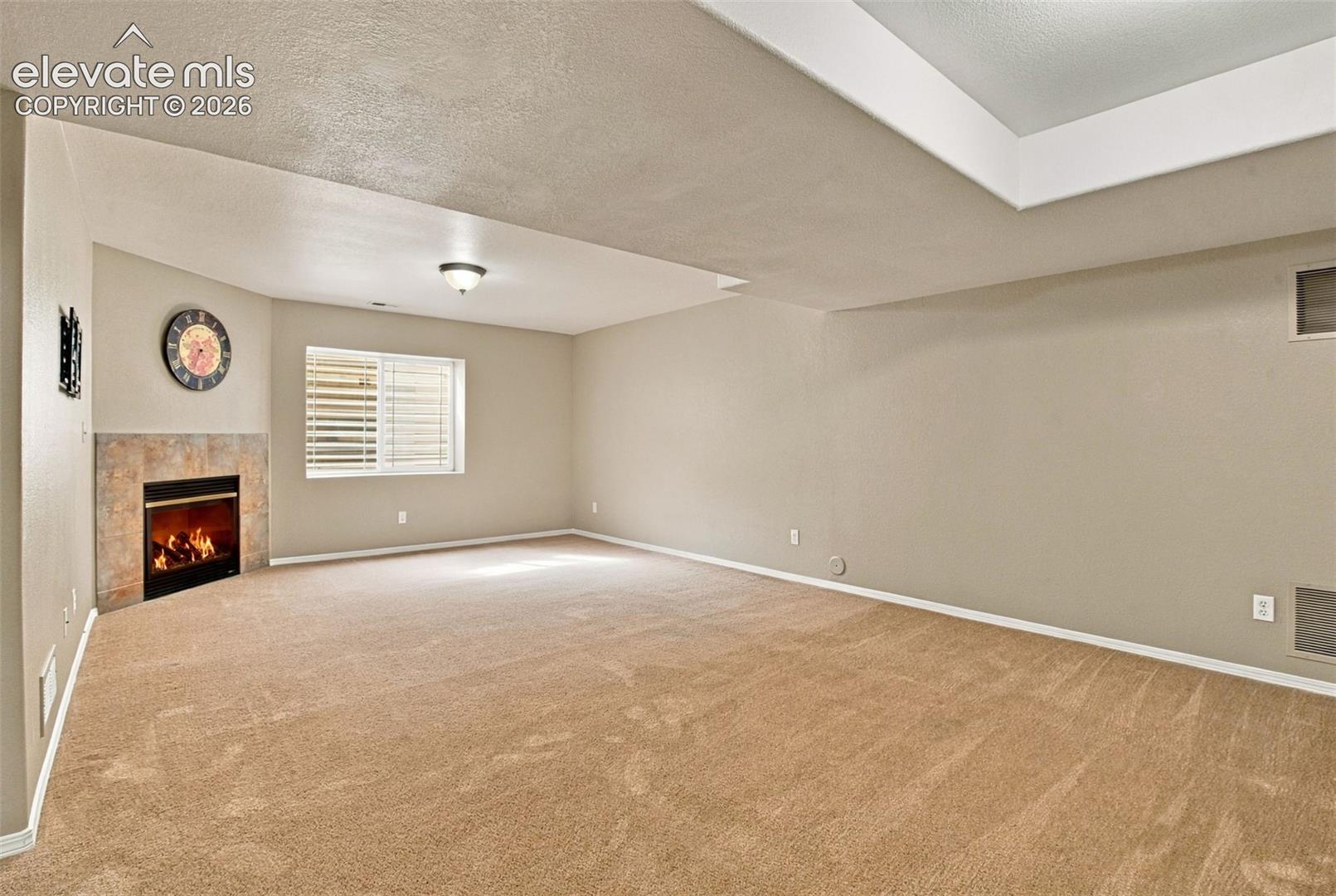 Spacious carpeted room featuring a corner fireplace with a tiled surround, a large window with white louvered shutters, and a tray ceiling detail
