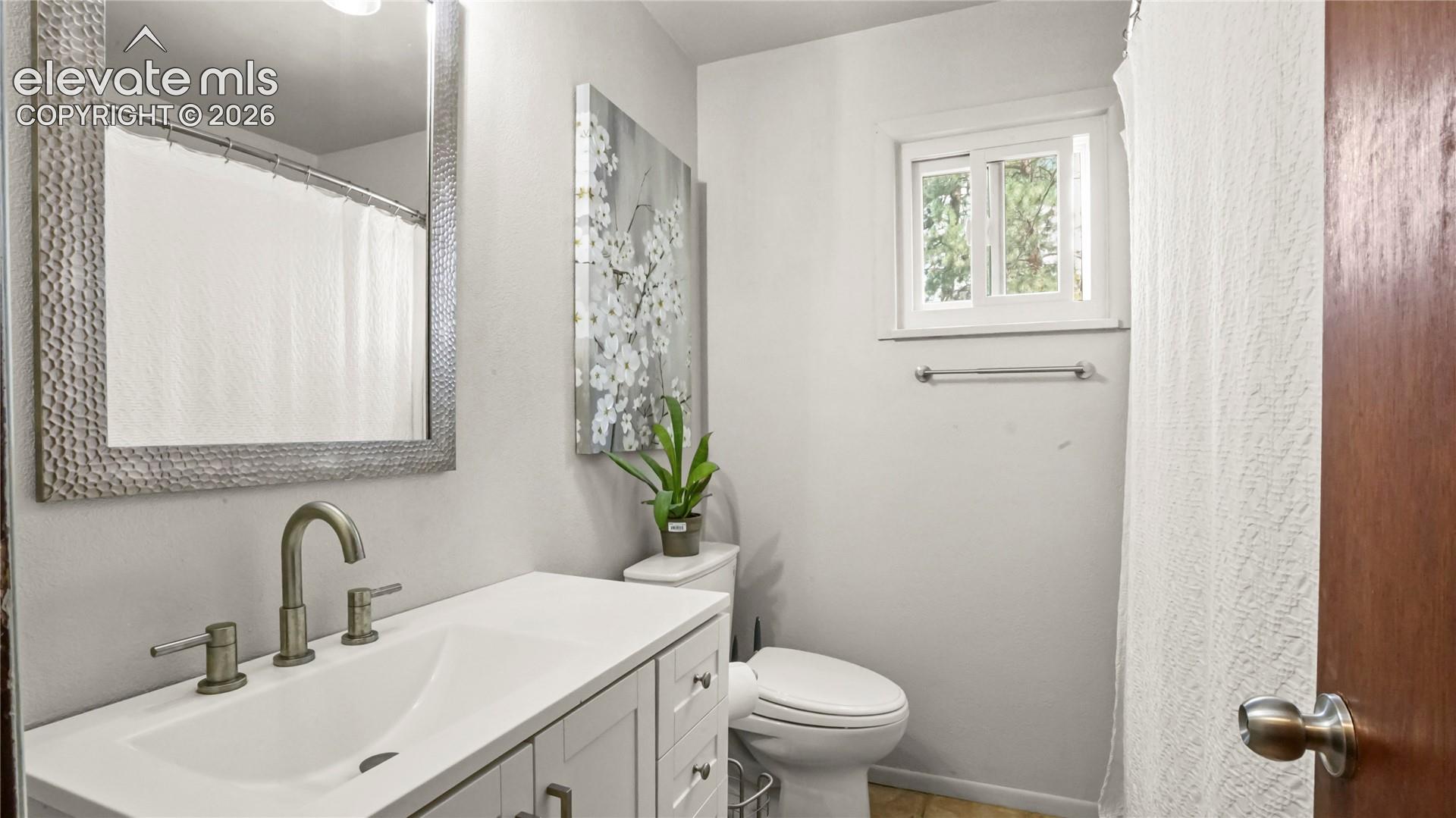 Stone countertop vanity with integrated sink, brushed nickel faucet, large framed mirror, white window frame, and a white toilet