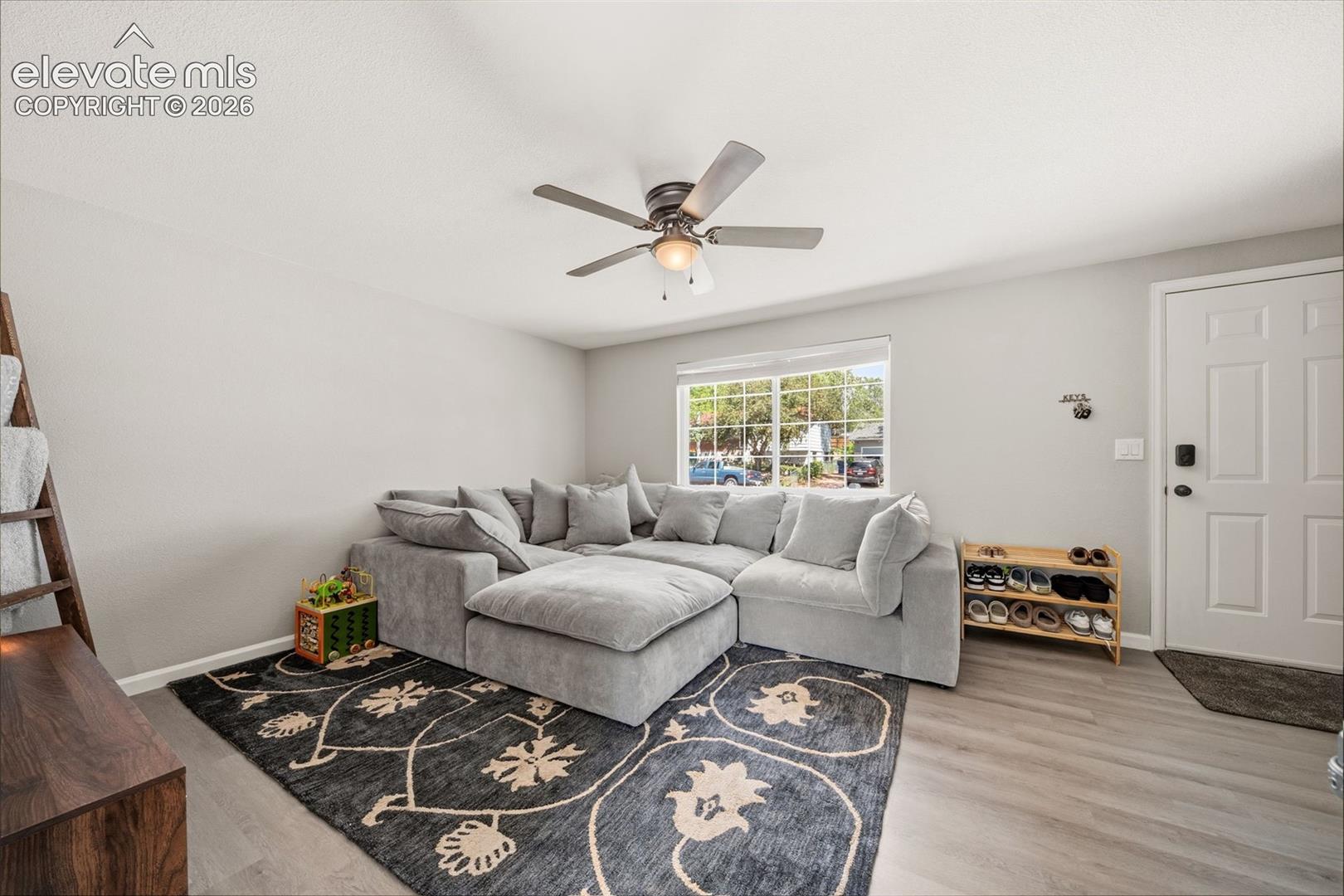 Wood-finish flooring, ceiling fan with integrated lighting, natural light from a large window, light-toned walls, and a paneled entry door