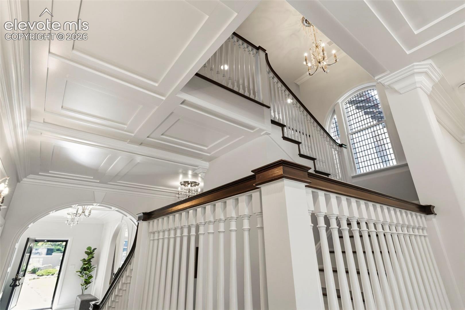 Foyer featuring a coffered ceiling, staircase with stained wood handrails and white balusters, an arched entry, and multi-light chandeliers