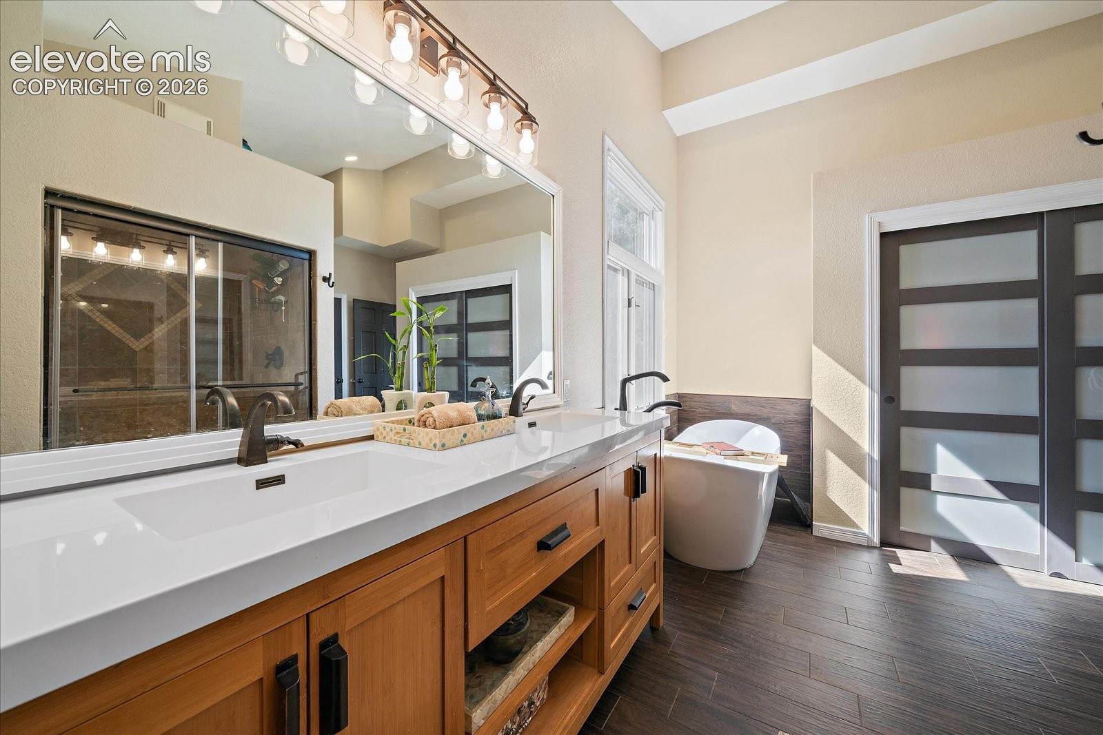 Dual-sink vanity with white surface, wood-finish cabinetry, freestanding soaking tub, wood-finish flooring, and frosted glass sliding door