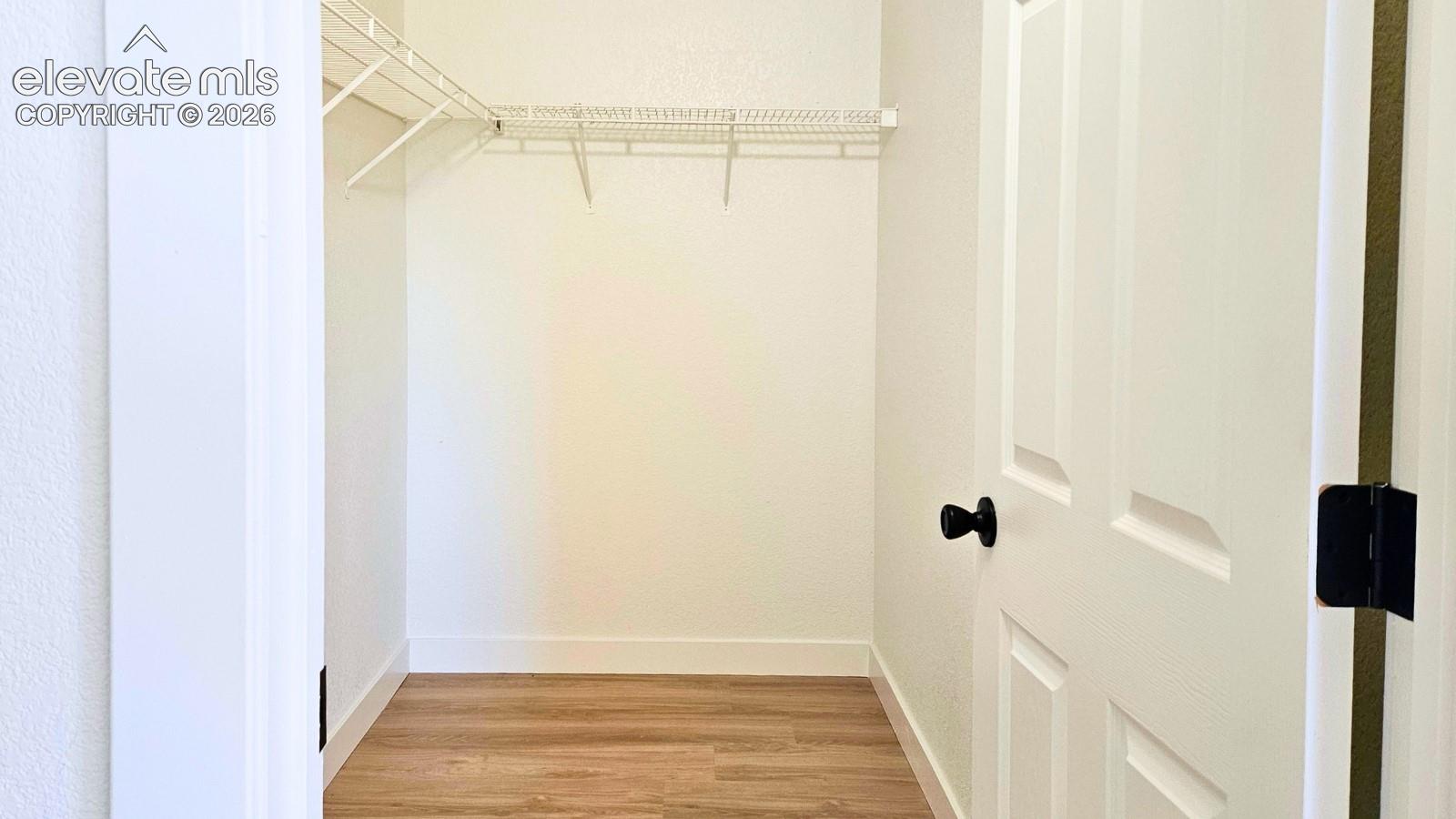 Storage space featuring wood-finish flooring, white walls, and built-in white wire shelving, accessed by a six-panel door with matte black hardware