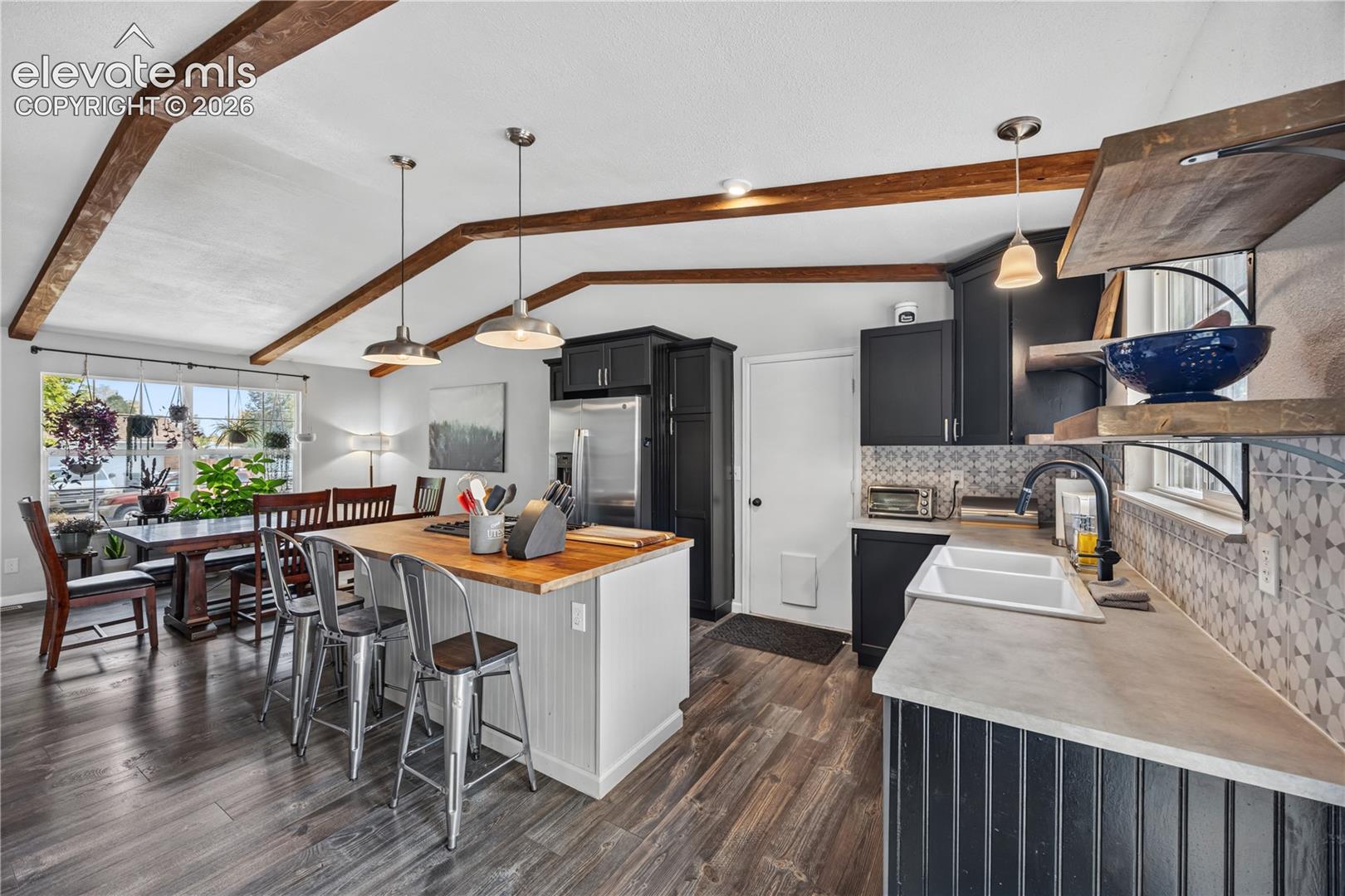 Vaulted ceiling with exposed wood beams, kitchen island with a wood countertop, dark-finish cabinetry, geometric tile backsplash, and wood-finish flooring