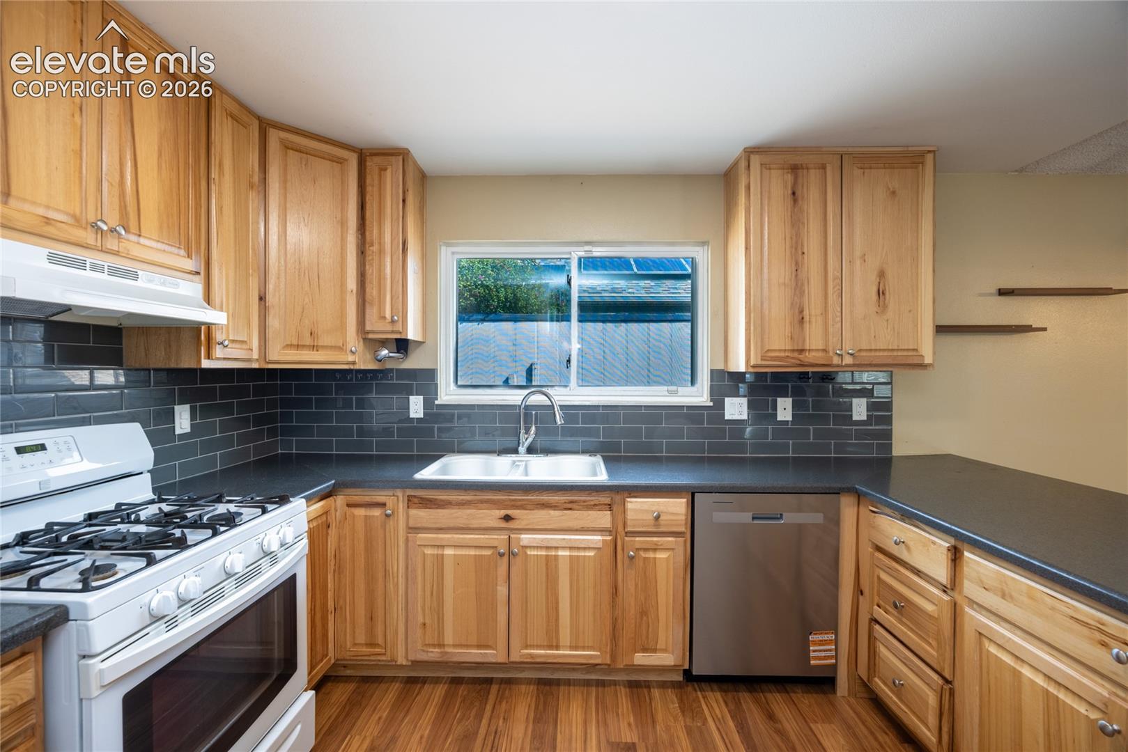 Wood-finish flooring, natural wood cabinetry, dark subway tile backsplash, stainless steel dishwasher, and black countertops