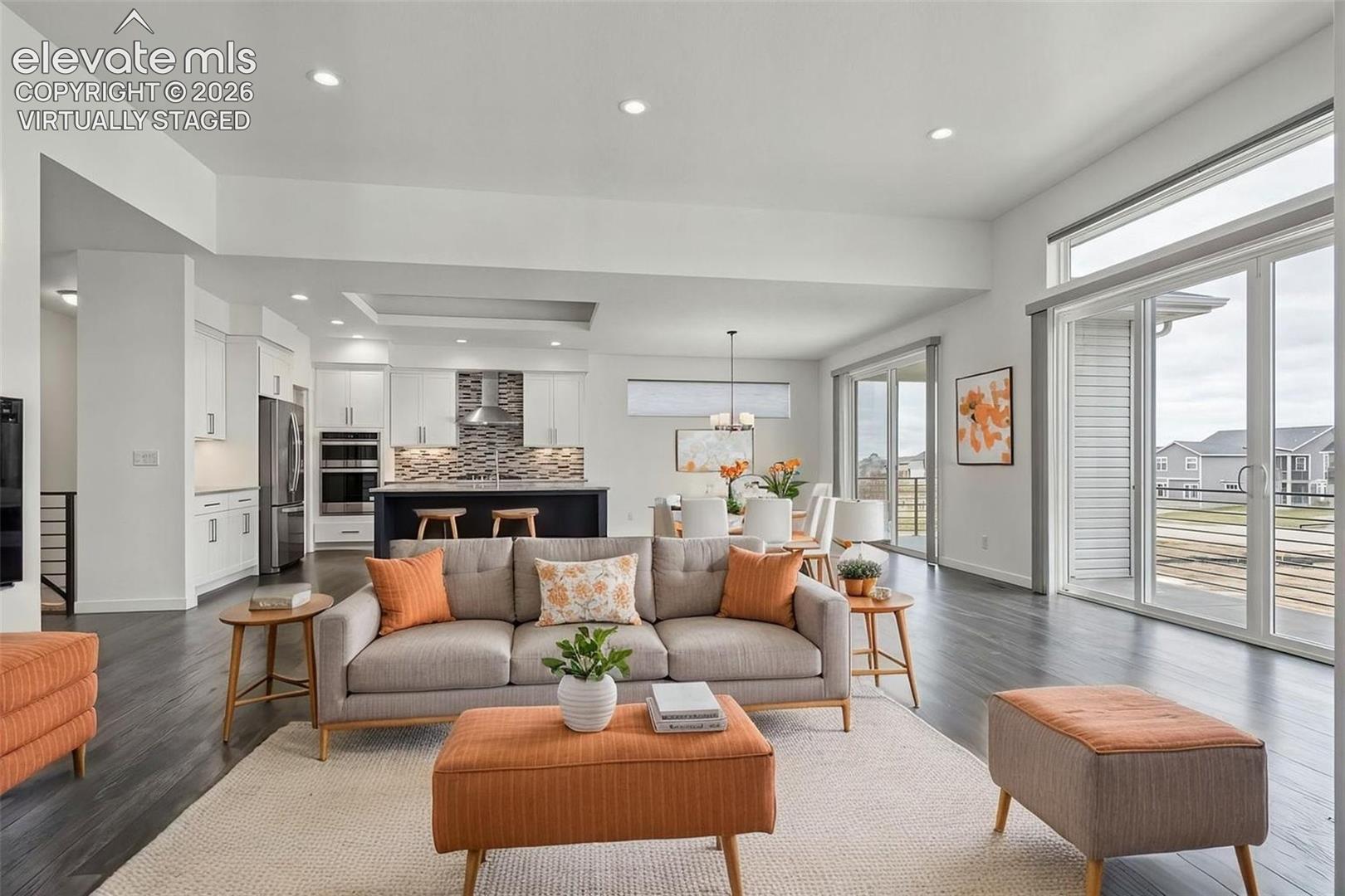 Open-concept living and kitchen area featuring recessed ceiling lighting, stainless steel appliances, a center island with seating, wood-finish flooring throughout, and sliding glass doors opening to a covered outdoor terrace with scenic vistas.  Virtually Staged