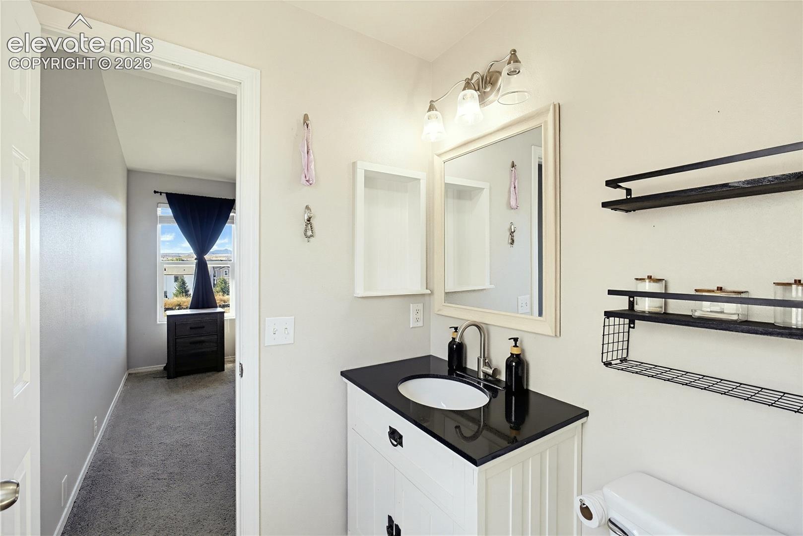 Vanity with a dark countertop and undermount sink, framed mirror with sconce lighting, and wall-mounted shelving