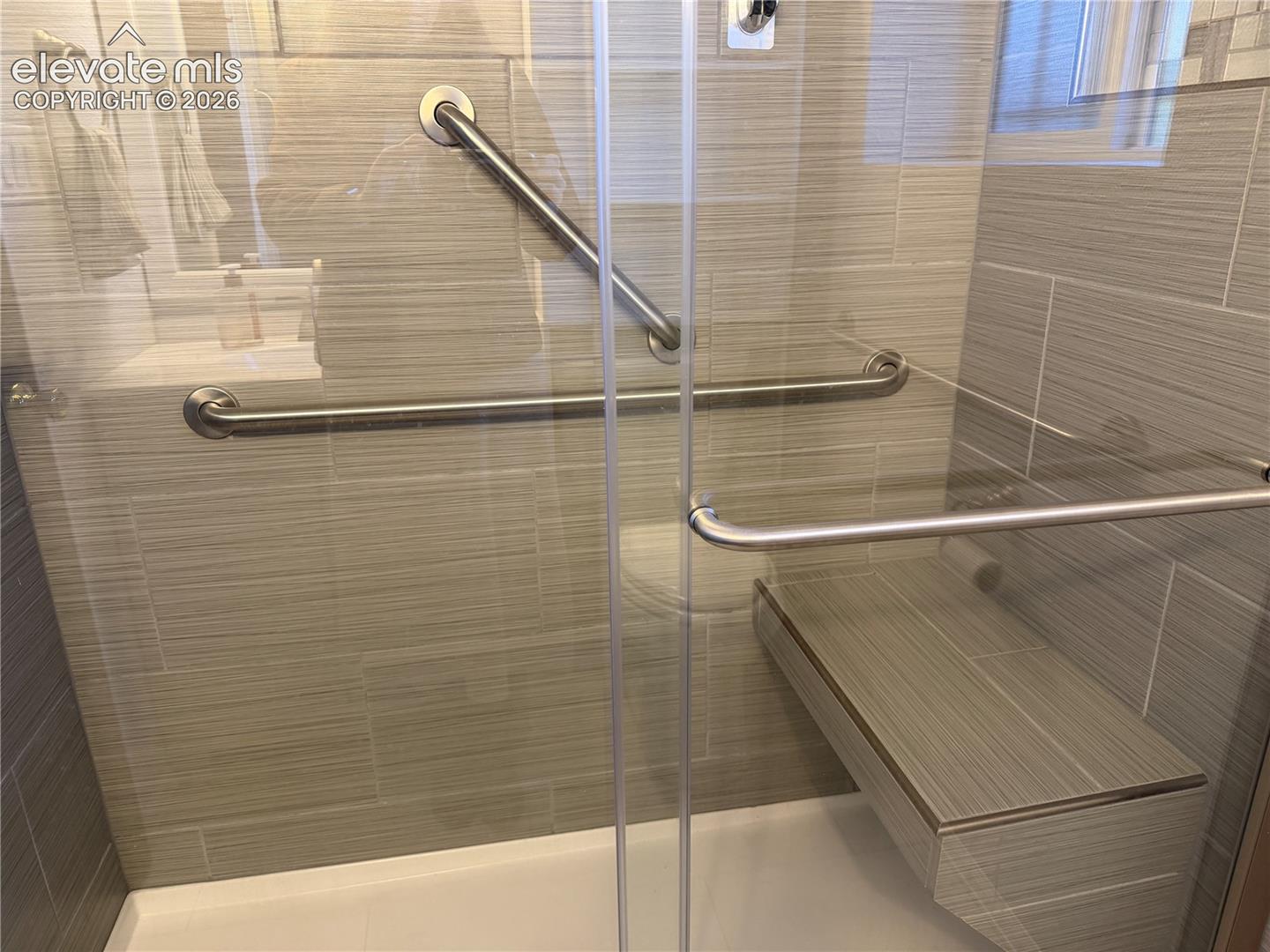 Updated bathroom in lower level with walk-in shower featuring textured wall tile, glass enclosure with brushed metal hardware, built-in tiled bench, and wall-mounted metal support bars