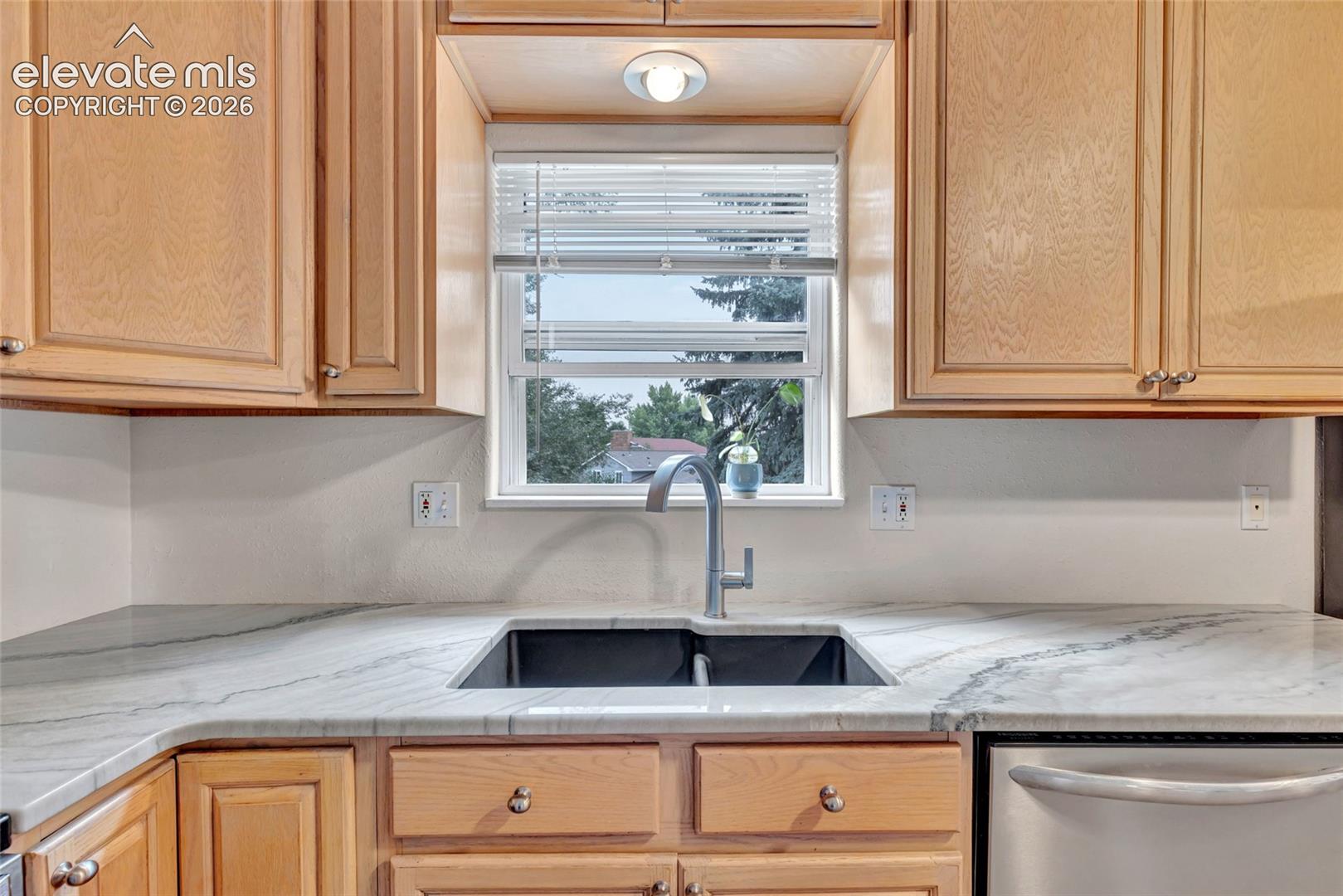 Kitchen with wood cabinetry, light-toned countertops, undermount sink with gooseneck faucet, and stainless steel dishwasher