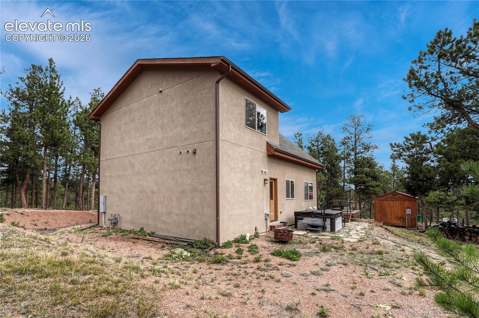 Stucco exterior featuring a pitched roofline, stone patio,  hot tub, and detached wood-frame outbuilding, all situated on a wooded lot