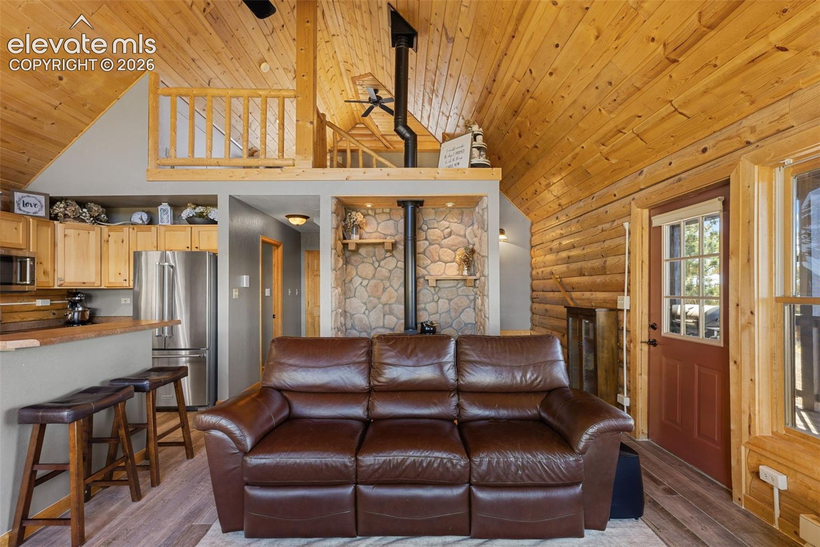 Vaulted wood-paneled ceiling, exposed log walls, floor-to-ceiling river rock hearth with a metal stove, and an elevated wooden loft area