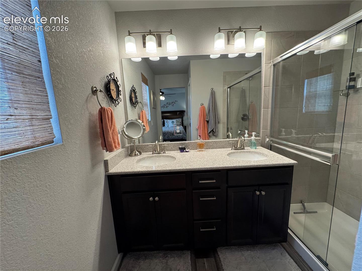 Double vanity featuring dark wood-finish cabinetry, a light-toned solid surface countertop with dual drop-in sinks, polished metal fixtures, and a glass-enclosed shower