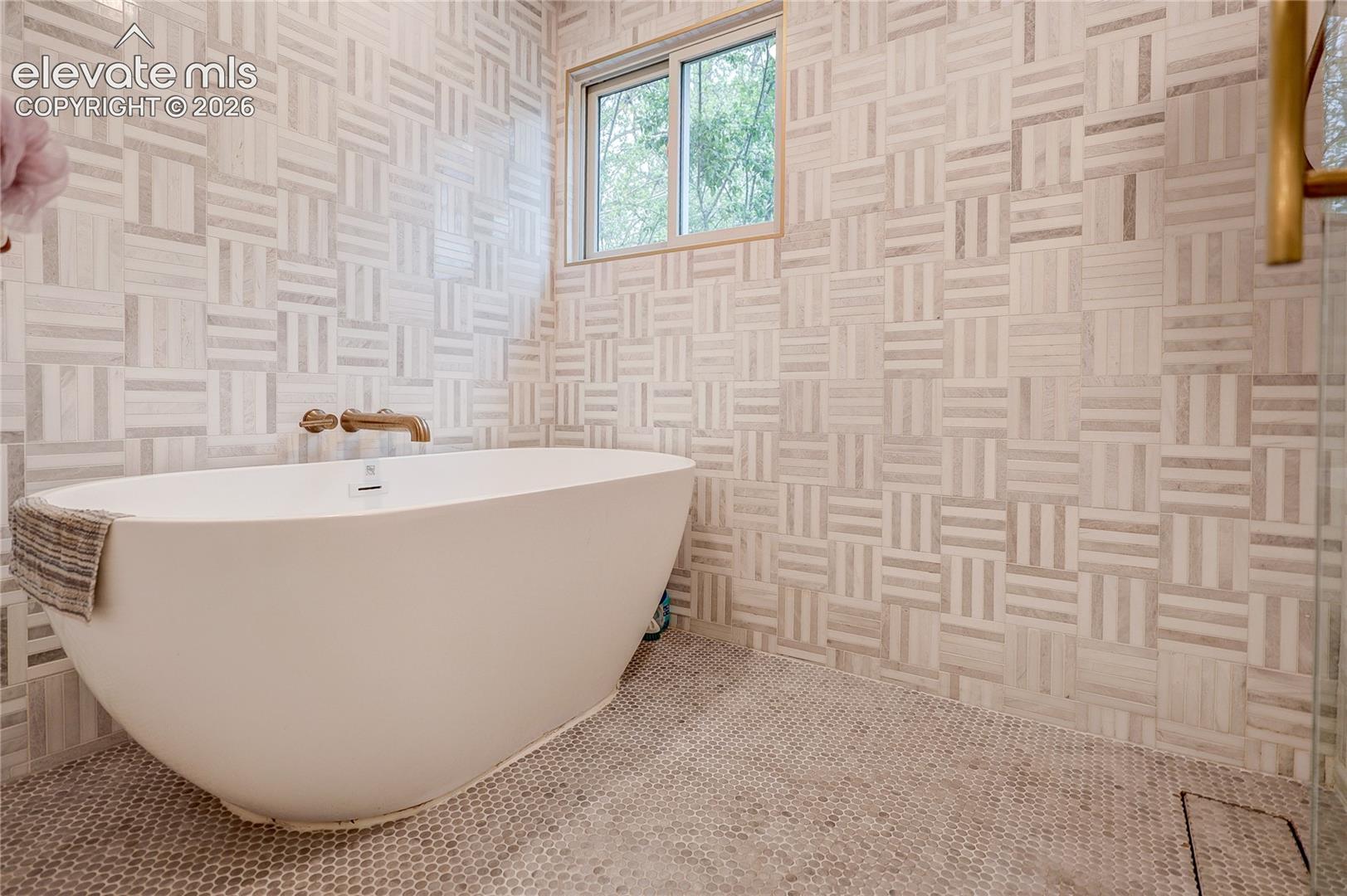 Freestanding soaking tub with wall-mounted bronze-finish faucet, surrounded by mosaic tile walls and penny tile flooring