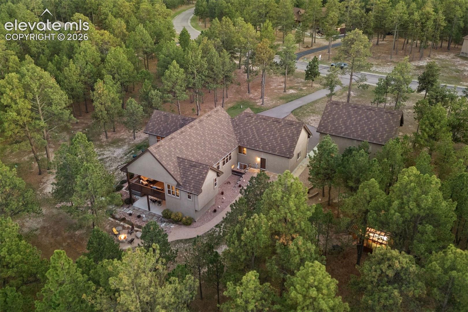 Aerial perspective of a multi-structure property featuring a pitched shingle roof, paved driveway, stone-paver hardscaping, and surrounding mature pine forest