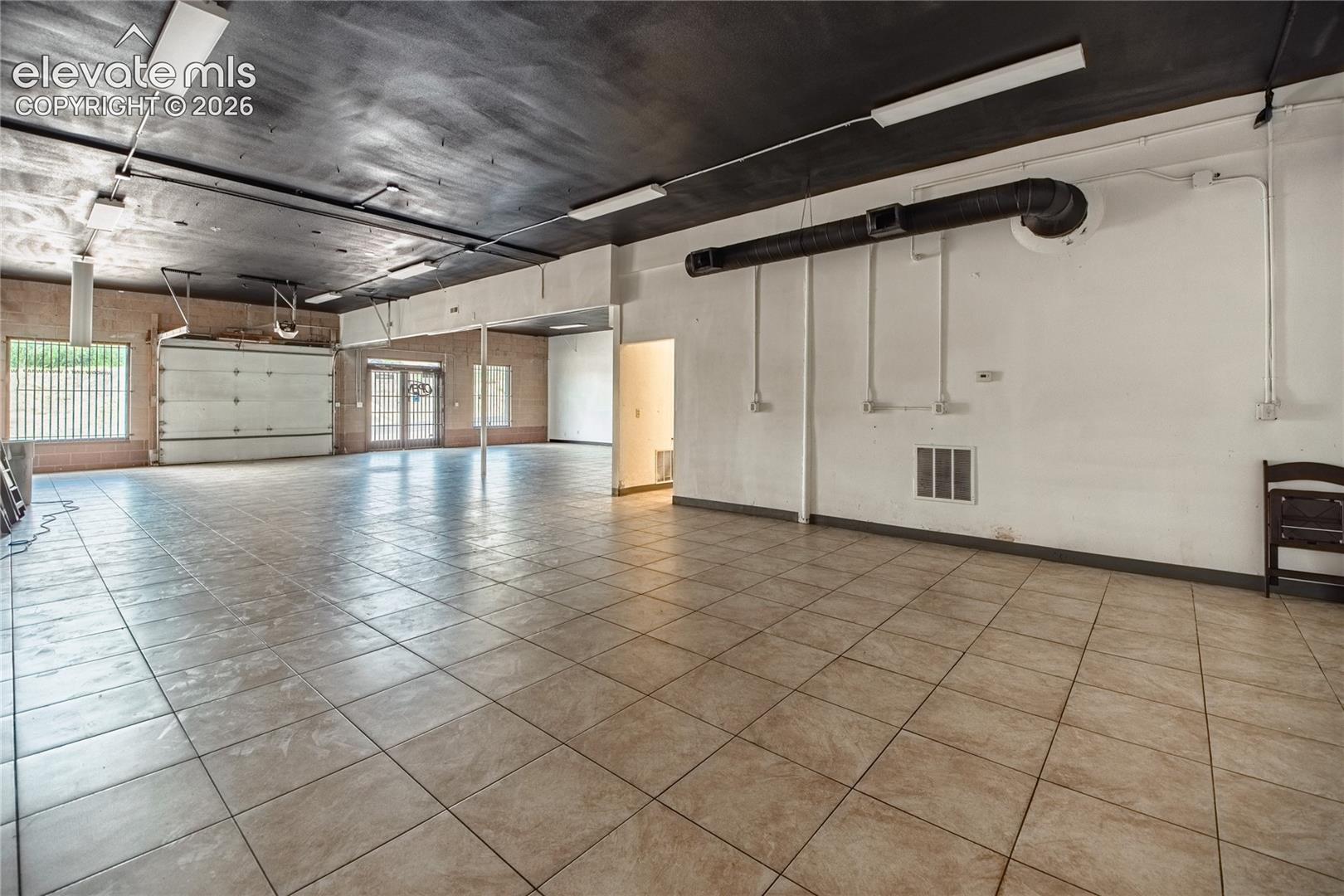 Expansive open-concept space featuring large format tile flooring, roll-up garage door, white walls, and exposed ceiling ductwork