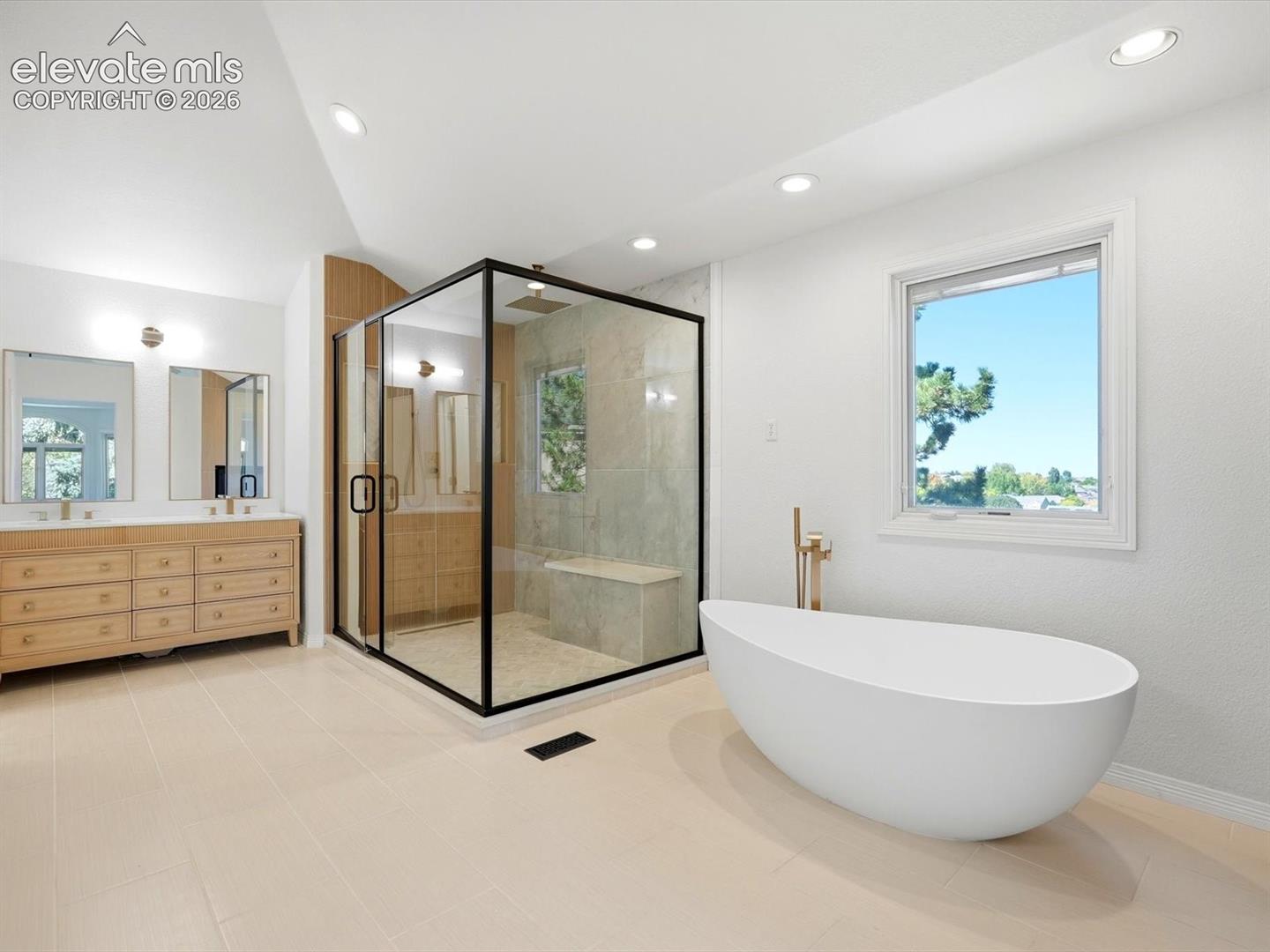 Freestanding soaking tub with floor-mounted brass faucet, walk-in glass-enclosed shower with black metal frame and built-in bench, and light-toned wood vanity with dual rectangular sinks