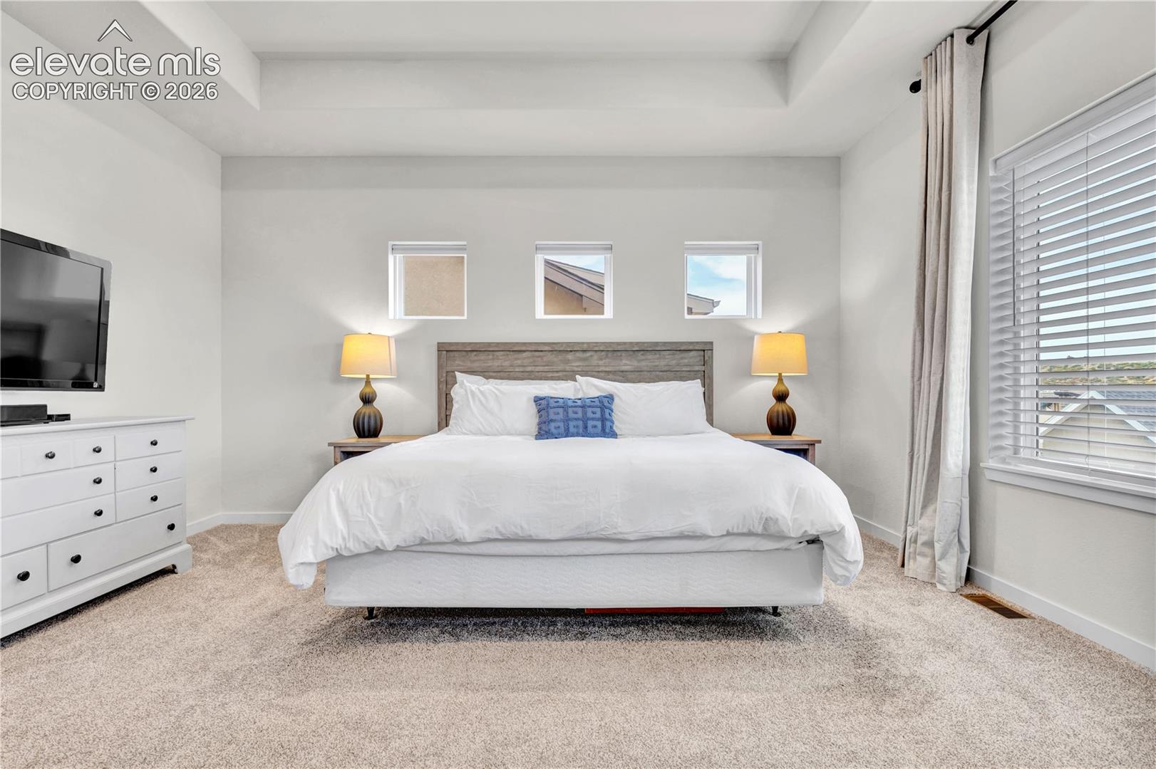 Tray ceiling with recessed lighting, large window with blinds, three upper windows, textured headboard, and carpet flooring