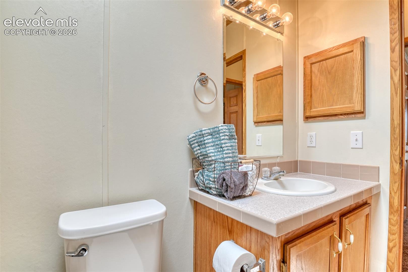 Bathroom featuring a white toilet, light wood-finish vanity with a tile countertop, integrated sink, and an overhead mirror with vanity lighting