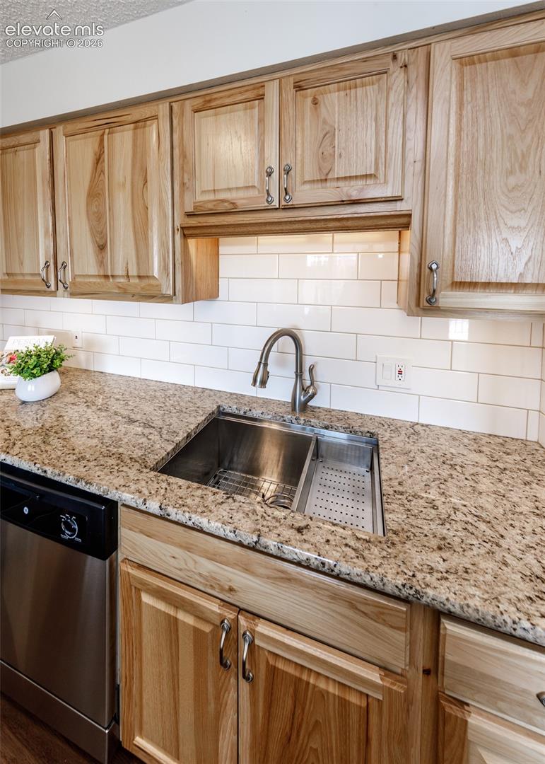 Kitchen featuring natural wood cabinetry, stone-finish countertops, white subway tile backsplash, stainless steel drop-in sink with pull-down faucet, and integrated stainless steel dishwasher