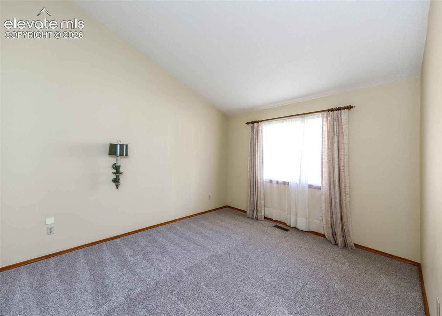 Vaulted ceiling, wall-mounted sconce, light-toned walls, window with trim, gray carpeting
